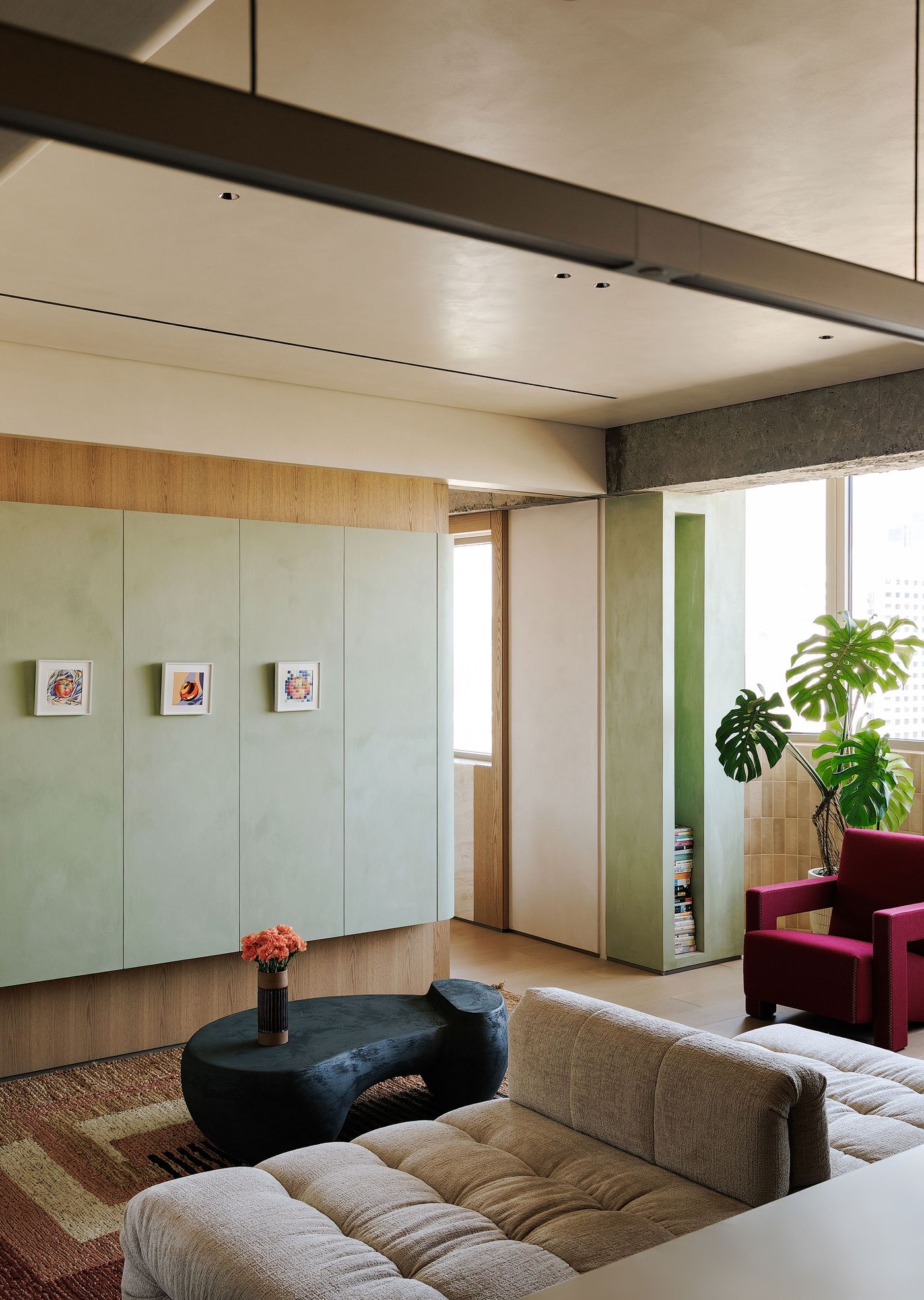 A multi-functional living area featuring a distinctive curved storage unit in a pale sage green hue, adorned with three small framed artworks. A plush cream modular sofa sits on a richly textured rug, while a sculptural dark gray coffee table adds a contemporary touch. Beyond, a vibrant red armchair hints at a more expansive living area, blending comfort with artistic expression. A multi-functional living area featuring a distinctive curved storage unit in a pale sage green hue, adorned with three small framed artworks. A plush cream modular sofa sits on a richly textured rug, while a sculptural dark gray coffee table adds a contemporary touch. Beyond, a vibrant red armchair hints at a more expansive living area, blending comfort with artistic expression.