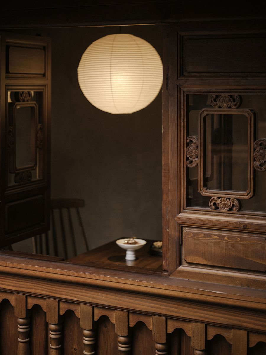 A softly glowing paper lantern hangs above a wooden table set with dishes, framed by ornate wooden windows and walls. A softly glowing paper lantern hangs above a wooden table set with dishes, framed by ornate wooden windows and walls.