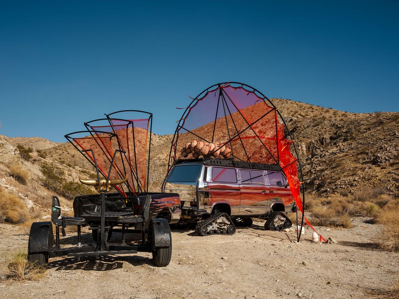 A silver van with red fabric canopies in a desert, alongside a unique trailer featuring decorative horns and unique wheel designs.