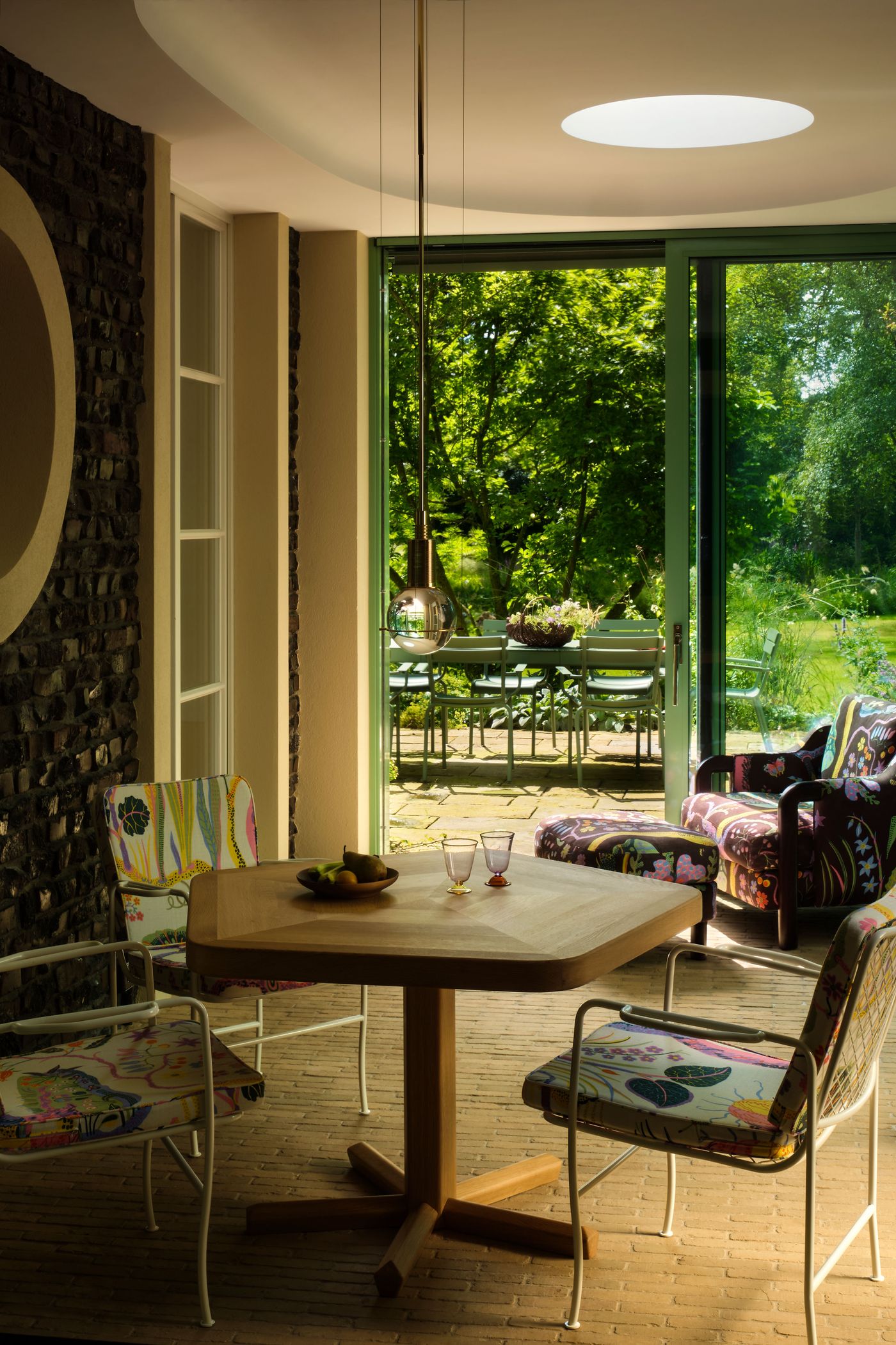 Bright dining area featuring a wooden table, colorful chairs, and lush garden view through open glass doors. Sunlight filters indoors.