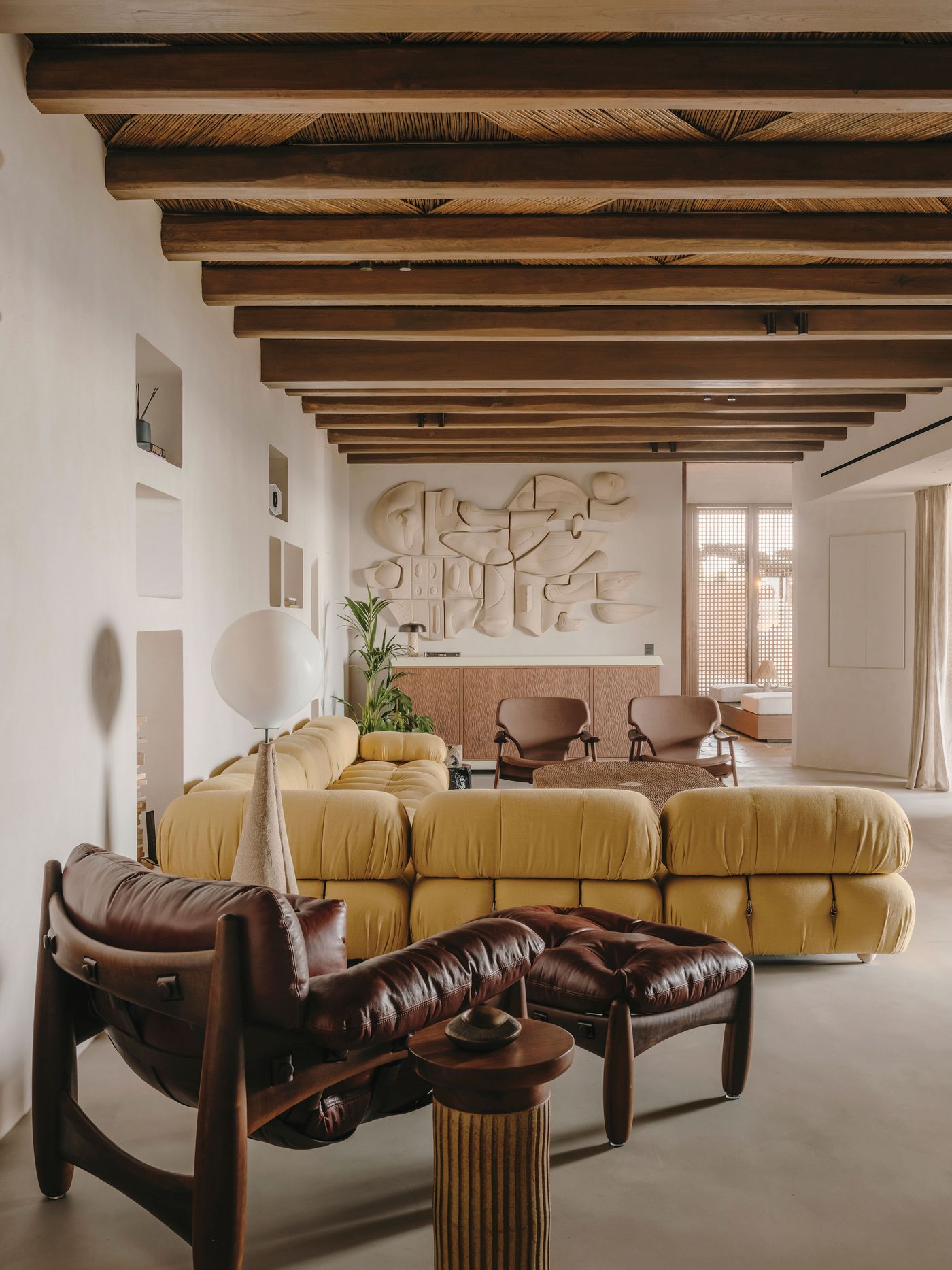Modern-meets-vernacular living room featuring a yellow sectional, brown leather chair, sculptural wall art, and warm wooden beams. Modern-meets-vernacular living room featuring a yellow sectional, brown leather chair, sculptural wall art, and warm wooden beams.