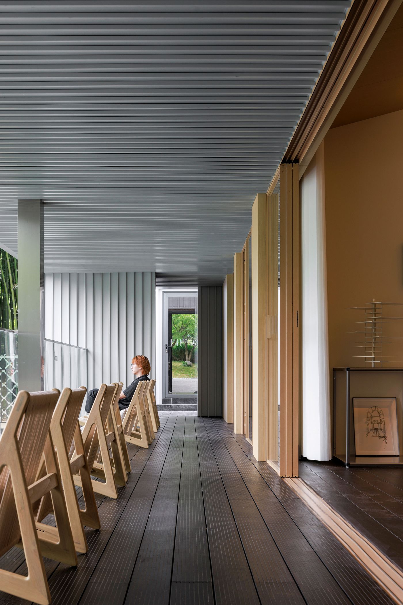 A long, linear perspective of a covered balcony at Wutopia Lab's Lake House, with a woman seated on one of several minimalist wooden folding chairs, looking towards the distant green outdoors. The space features a dark slatted floor and a light, corrugated ceiling, with a playful contrast between solid walls and a series of light wooden vertical slats that provide partial privacy and aesthetic interest. A long, linear perspective of a covered balcony at Wutopia Lab's Lake House, with a woman seated on one of several minimalist wooden folding chairs, looking towards the distant green outdoors. The space features a dark slatted floor and a light, corrugated ceiling, with a playful contrast between solid walls and a series of light wooden vertical slats that provide partial privacy and aesthetic interest.
