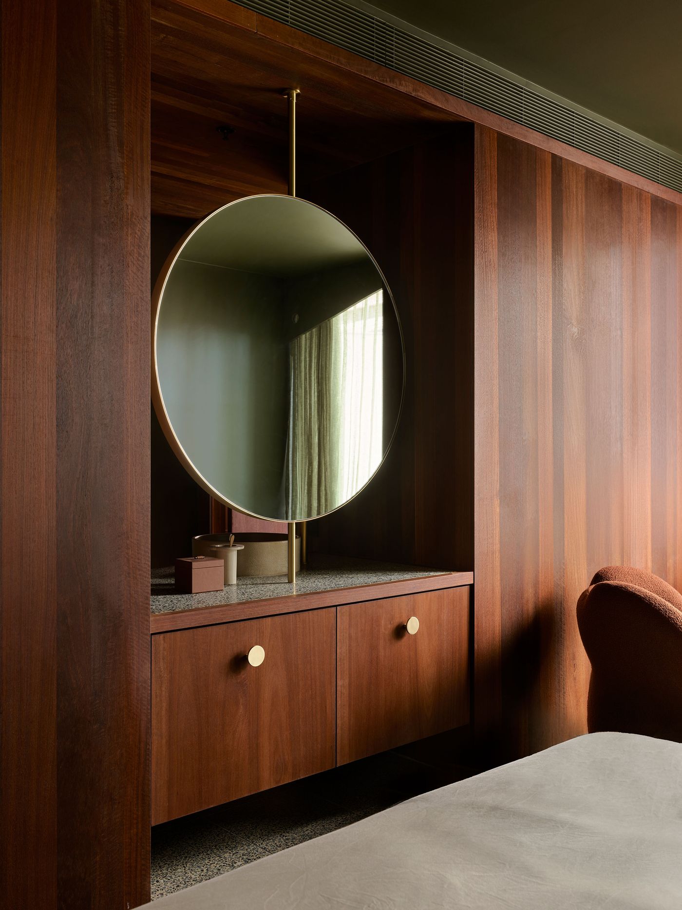 A round mirror hangs above a stylish wooden dresser with gold knobs, set against a backdrop of rich wood paneling and soft light.