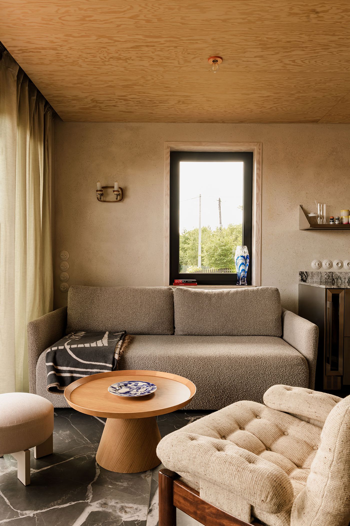 Cozy interior featuring a textured gray sofa, wooden coffee table with a decorative plate, and a view through the window framed by plants. Cozy interior featuring a textured gray sofa, wooden coffee table with a decorative plate, and a view through the window framed by plants.
