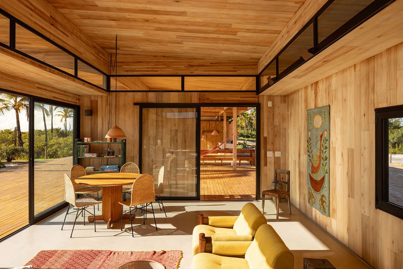 A sun-drenched living and dining area, clad in warm timber, features a circular dining table with wicker chairs and a vibrant yellow sofa. Expansive glass doors frame views of the outdoor deck and tropical foliage, embodying a light-filled, modern rustic aesthetic with bohemian touches.