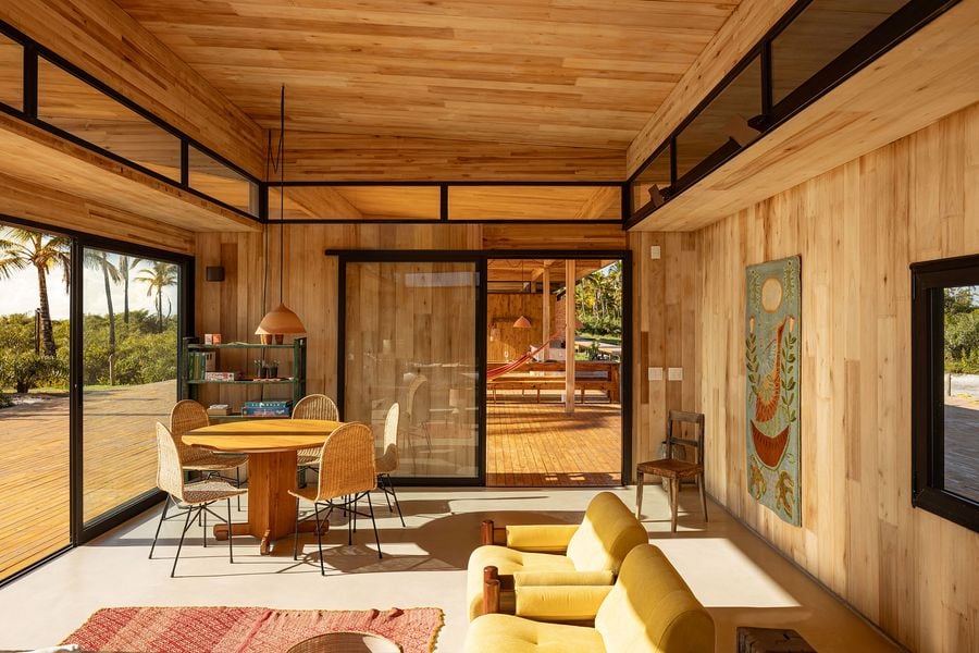 A sun-drenched living and dining area, clad in warm timber, features a circular dining table with wicker chairs and a vibrant yellow sofa. Expansive glass doors frame views of the outdoor deck and tropical foliage, embodying a light-filled, modern rustic aesthetic with bohemian touches.