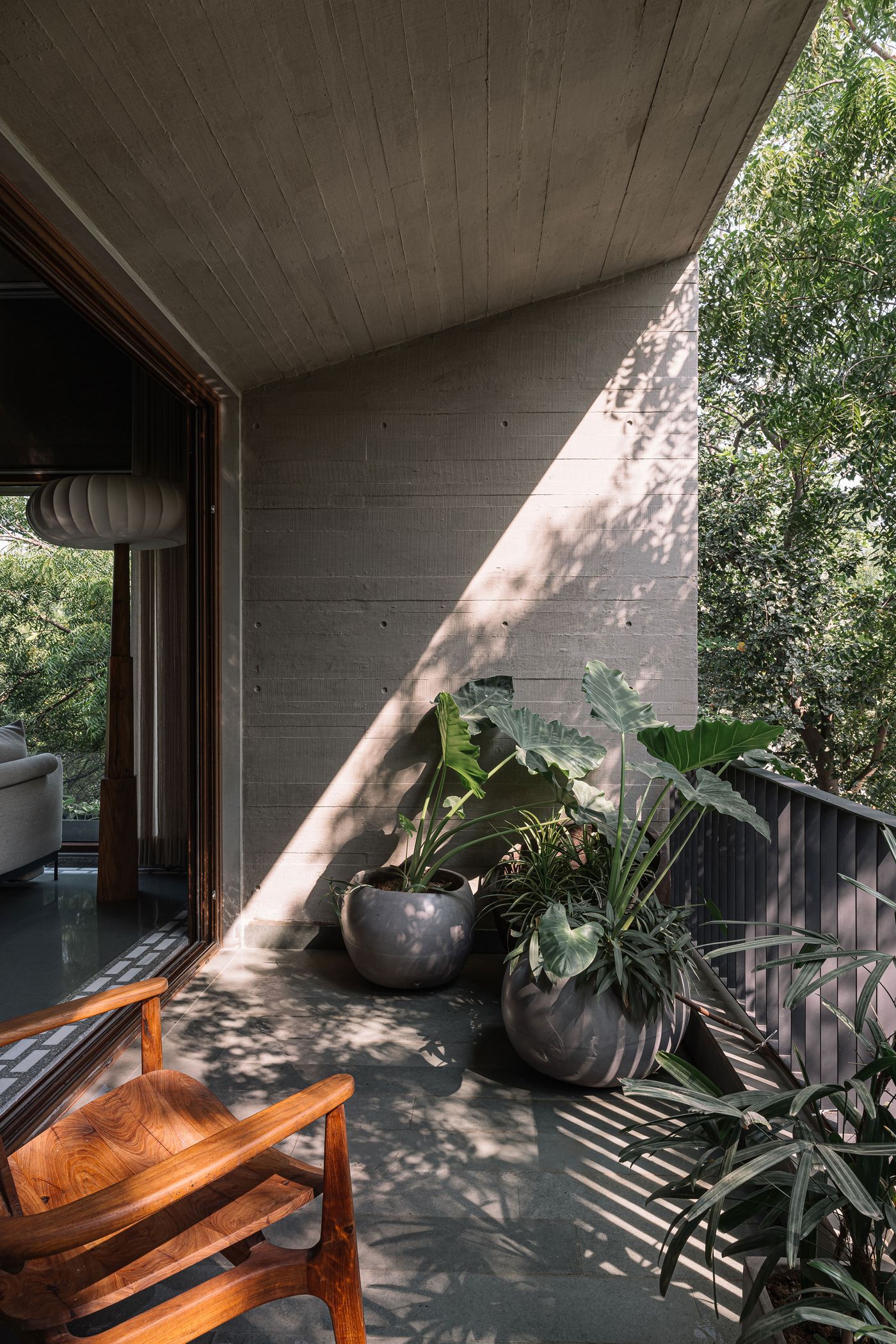 A serene outdoor balcony area, seamlessly extending from an indoor space with a concrete ceiling and wall. Large potted plants introduce lush greenery, enhancing the biophilic design. Sunlight streams in, casting dappled shadows through the surrounding trees, creating a peaceful retreat with a natural, rustic ambiance and a comfortable wooden chair.