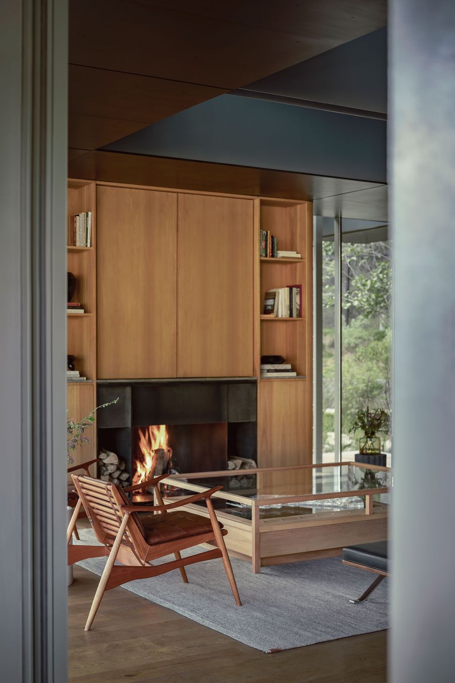 Cozy living room with a wooden bookshelf, a glass coffee table, and a warm fire glowing in the fireplace, seen through a doorway.