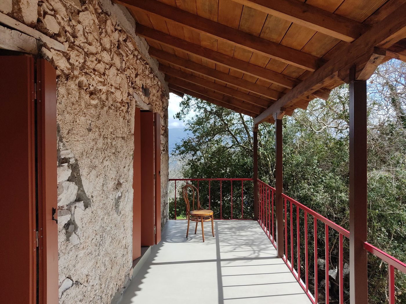 Traditional timber beam construction creates geometric shadows across the upper-level terrace, where coral-red railings provide contemporary safety elements. A solitary bentwood chair anchors the space, emphasizing the project's balance between historical preservation and modern comfort within the Mediterranean landscape.