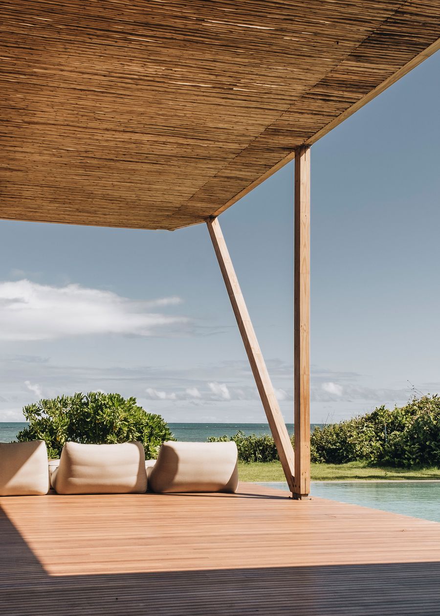 A serene outdoor space with wooden decking, minimal beige cushions, and a stunning ocean view under a bamboo roof.