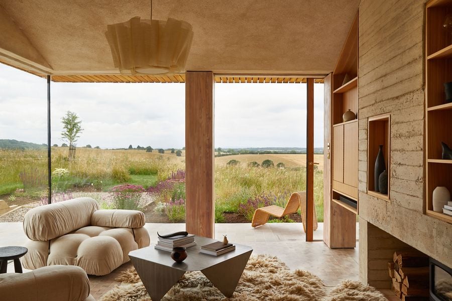 The living space of The Maker's Barn, a biophilic design by Hutch Design, offers an immersive connection to nature. Expansive glass walls frame a sweeping vista of golden fields and wildflowers. Inside, plush, cream-colored modular seating and a unique sculptural coffee table rest on a shaggy rug, complemented by a large, flowing fabric light fixture and a striking wooden rocking chair that blends rustic charm with modern minimalism.