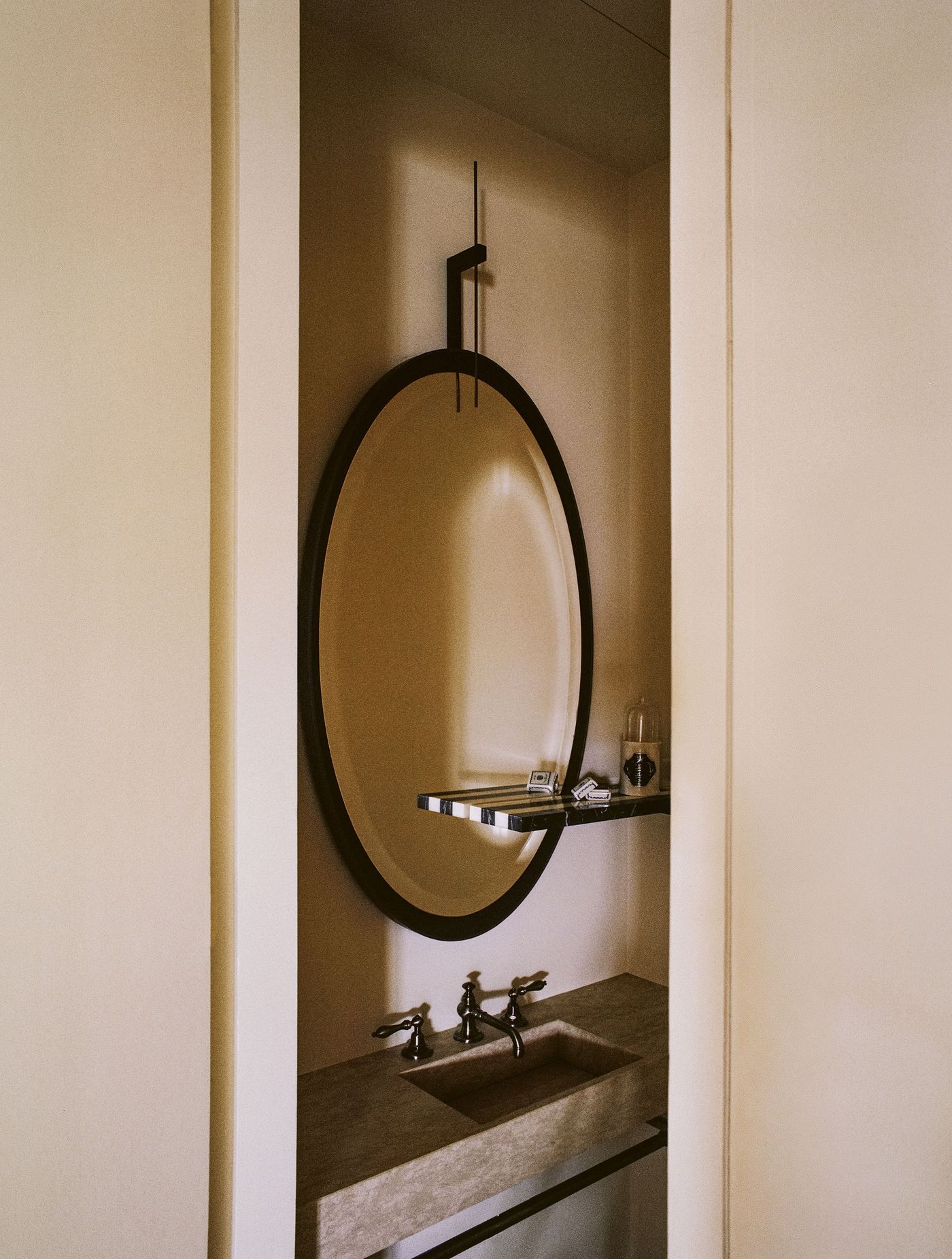 A narrow view into a minimalist powder room, featuring a large, elegantly framed oval mirror suspended from the ceiling. Below, a sleek, rectangular basin in a light stone material with exposed brass taps adds a touch of understated luxury, reflecting a refined aesthetic. A narrow view into a minimalist powder room, featuring a large, elegantly framed oval mirror suspended from the ceiling. Below, a sleek, rectangular basin in a light stone material with exposed brass taps adds a touch of understated luxury, reflecting a refined aesthetic.