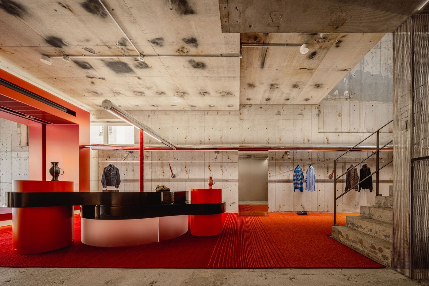 The ground floor combines an expansive red-orange surface with a sculptural display counter of glossy black and cylindrical forms. Exposed concrete walls and ceiling remain visibly patched and stained, while chrome rails, transparent elements and the adjacent raw staircase introduce a sharper, more precise layer into the Brutalist shell.