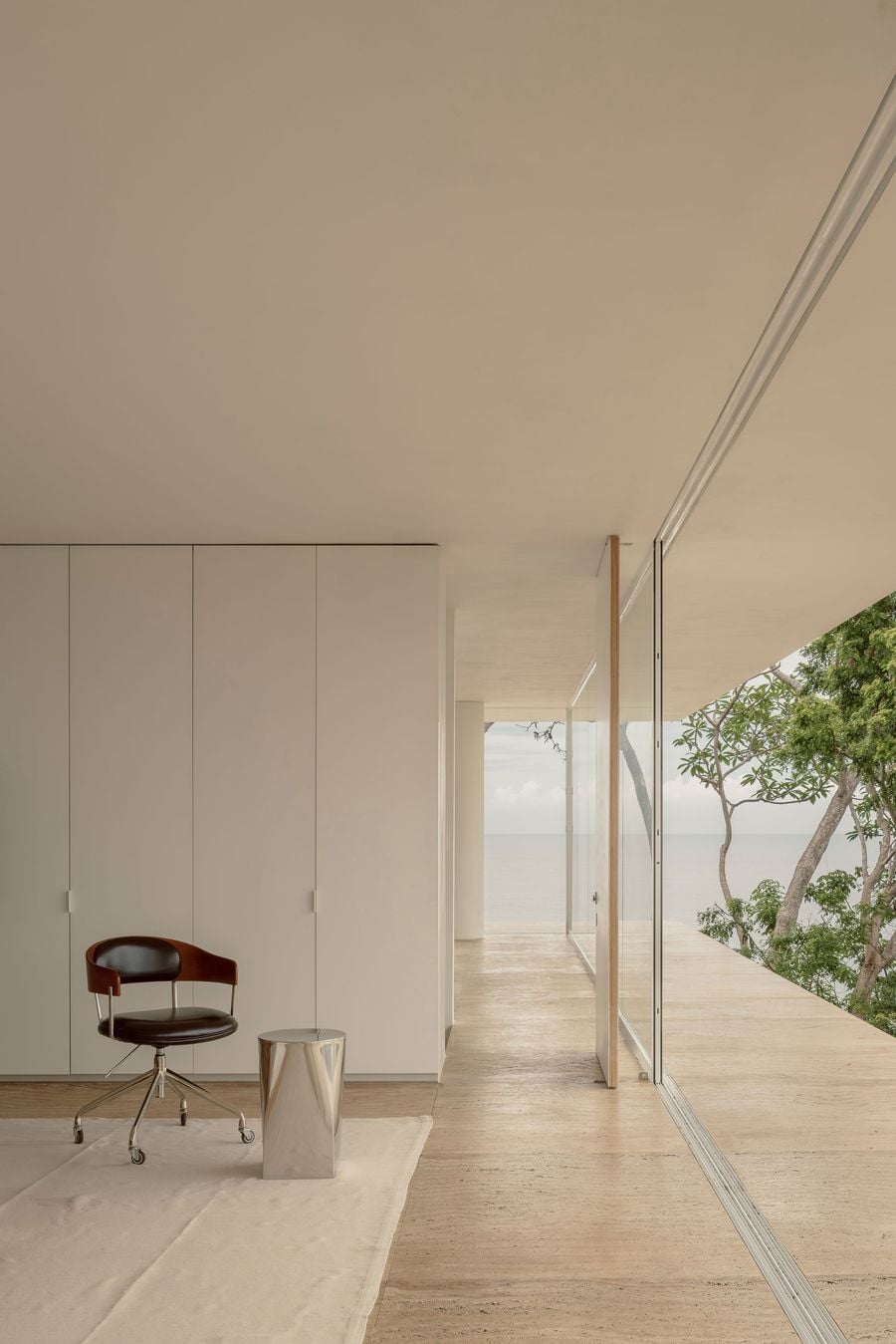 Flush white joinery, a leather desk chair and a mirror-polished side table compose a quiet corner of the bedroom, where the veined floor continues seamlessly onto the terrace. Beyond the sliding glass, slender trunks and a hazy sea horizon extend the room into the landscape. Flush white joinery, a leather desk chair and a mirror-polished side table compose a quiet corner of the bedroom, where the veined floor continues seamlessly onto the terrace. Beyond the sliding glass, slender trunks and a hazy sea horizon extend the room into the landscape.