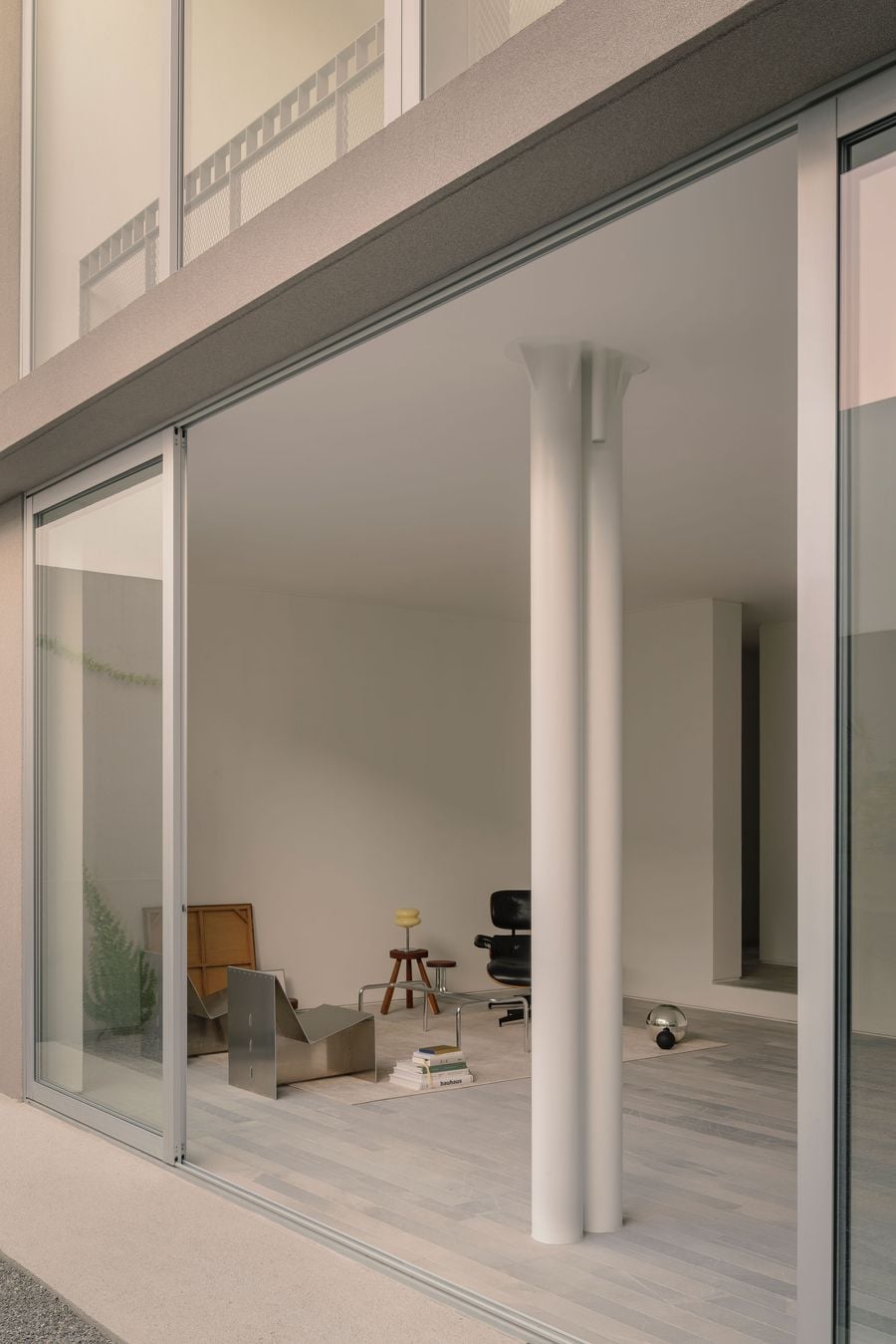 A close view through Casa Tlaloc’s floor-to-ceiling glazing highlights the delicate relationship between structure and interior. A cluster of slender white columns stands against the glass, framing a sparse living room furnished with sculptural metal, timber and black leather pieces, while reflections layer the space with the terrace beyond. A close view through Casa Tlaloc’s floor-to-ceiling glazing highlights the delicate relationship between structure and interior. A cluster of slender white columns stands against the glass, framing a sparse living room furnished with sculptural metal, timber and black leather pieces, while reflections layer the space with the terrace beyond.