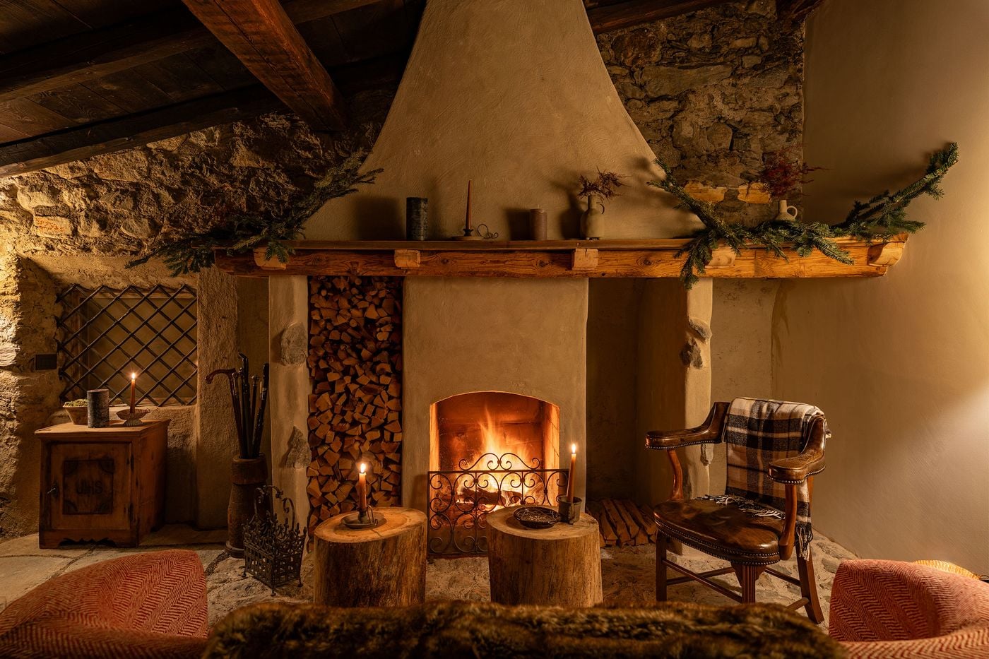 A firelit sitting room at Castel Badia gathers around a tapering plastered chimney hood with a rustic timber mantel draped in fir boughs. Rough stone walls, a niche of stacked logs, tree-stump side tables, a leather armchair with a tartan throw and an antique cabinet carved with an IHS monogram complete the scene. A firelit sitting room at Castel Badia gathers around a tapering plastered chimney hood with a rustic timber mantel draped in fir boughs. Rough stone walls, a niche of stacked logs, tree-stump side tables, a leather armchair with a tartan throw and an antique cabinet carved with an IHS monogram complete the scene.