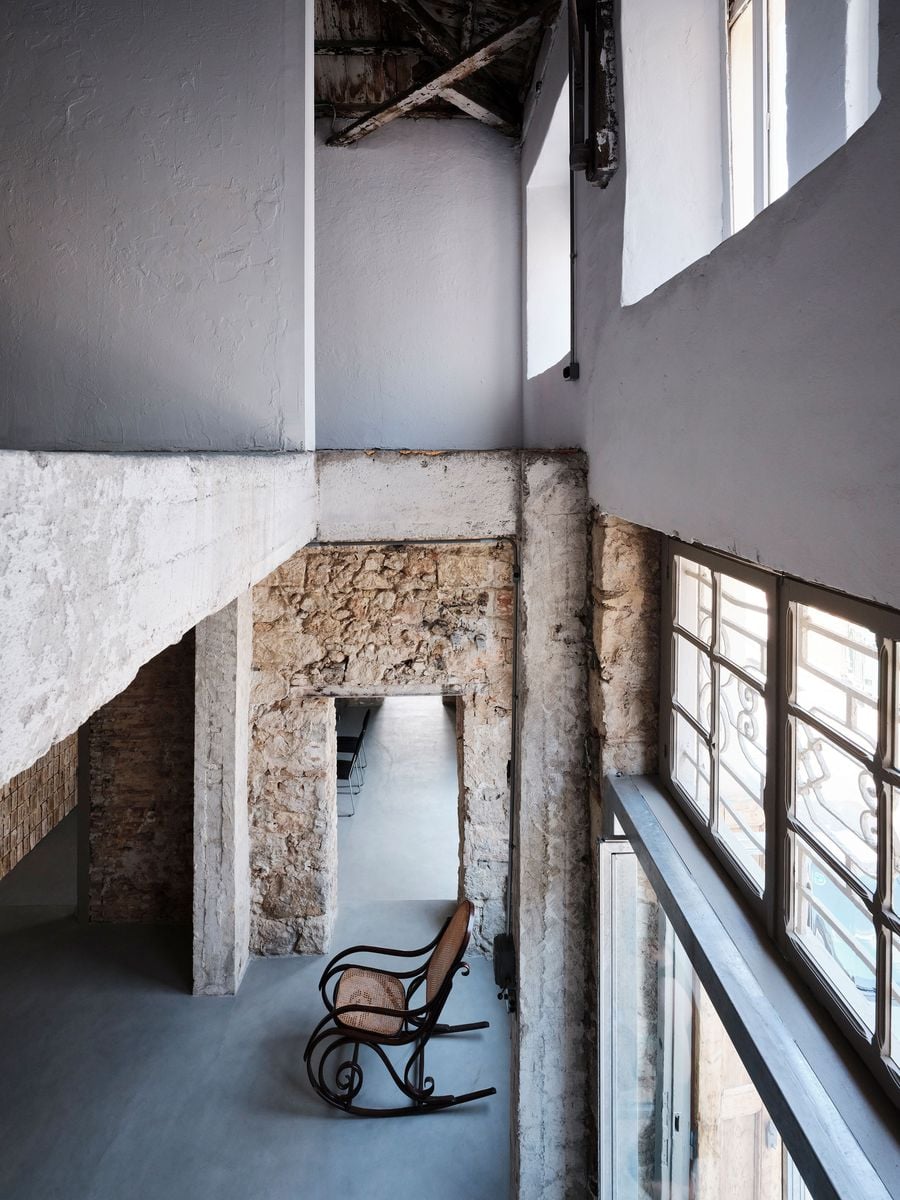 A high-level view reveals the building’s layered vertical organisation, with the concrete mezzanine cutting through the original masonry shell beneath the exposed timber roof. Below, a vintage cane rocking chair sits beside tall windows, while aligned openings establish long sightlines through the renovated ground floor. A high-level view reveals the building’s layered vertical organisation, with the concrete mezzanine cutting through the original masonry shell beneath the exposed timber roof. Below, a vintage cane rocking chair sits beside tall windows, while aligned openings establish long sightlines through the renovated ground floor.