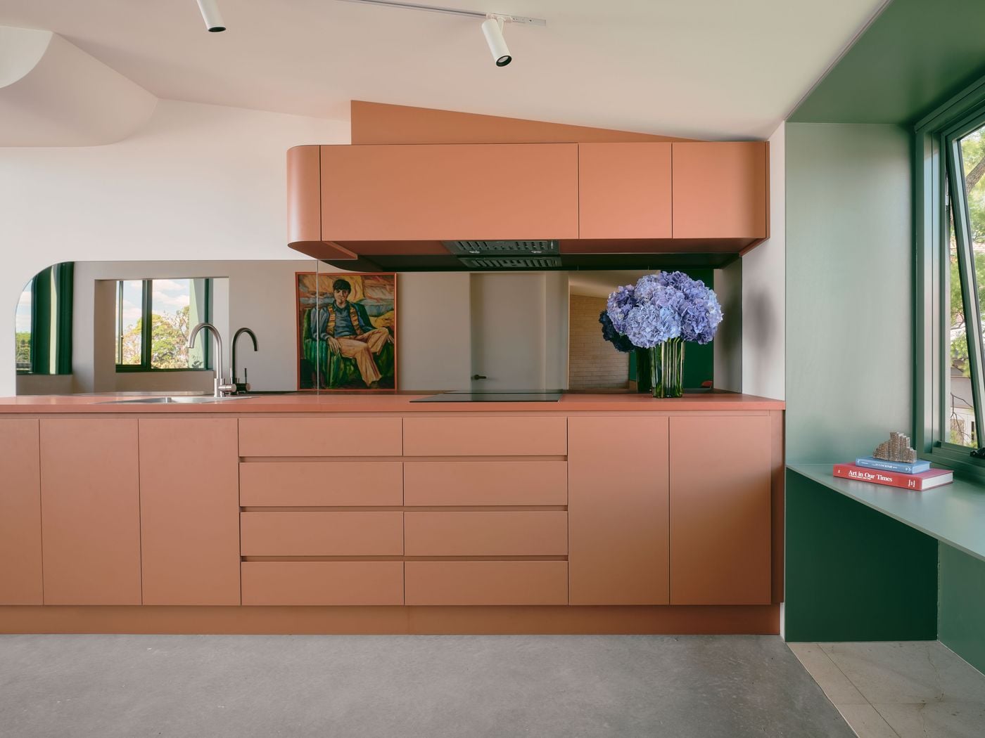Terracotta cabinetry gives Denj Lane’s compact kitchen a warm, graphic presence, its rounded upper corner continuing the project’s recurring curved geometry. A mirrored splashback visually expands the space and reflects artwork and flowers, while an adjoining sage-green window seat introduces a contrasting colour block and useful built-in surface. Terracotta cabinetry gives Denj Lane’s compact kitchen a warm, graphic presence, its rounded upper corner continuing the project’s recurring curved geometry. A mirrored splashback visually expands the space and reflects artwork and flowers, while an adjoining sage-green window seat introduces a contrasting colour block and useful built-in surface.