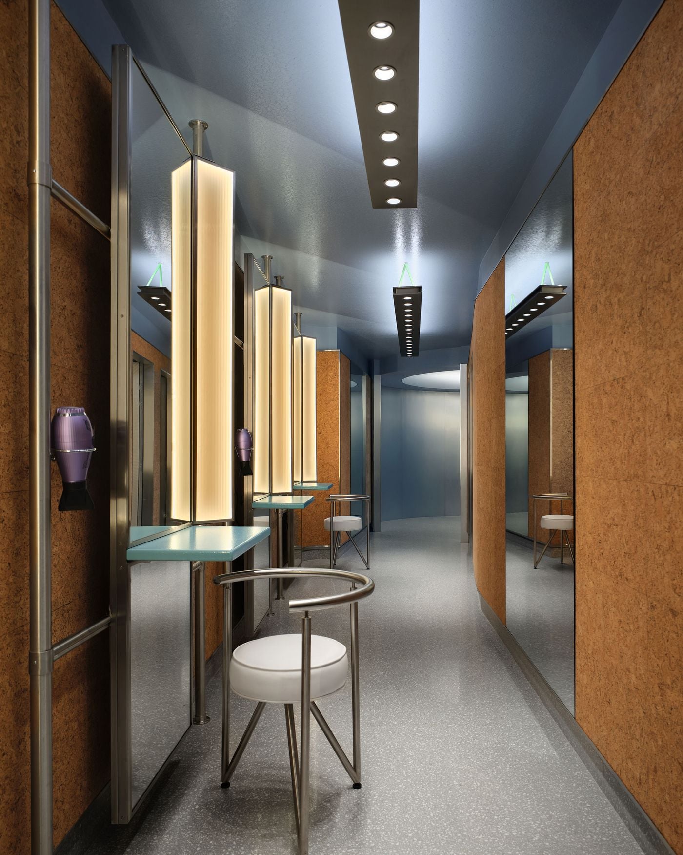 A grooming area in the women’s locker room combines cork-clad walls, illuminated vertical mirrors and slim stainless-steel furniture beneath a cool blue ceiling. Tubular metal frames and suspended linear lights give the space a crisp, retro-futurist character, softened by pale blue counters and white upholstered stools. A grooming area in the women’s locker room combines cork-clad walls, illuminated vertical mirrors and slim stainless-steel furniture beneath a cool blue ceiling. Tubular metal frames and suspended linear lights give the space a crisp, retro-futurist character, softened by pale blue counters and white upholstered stools.