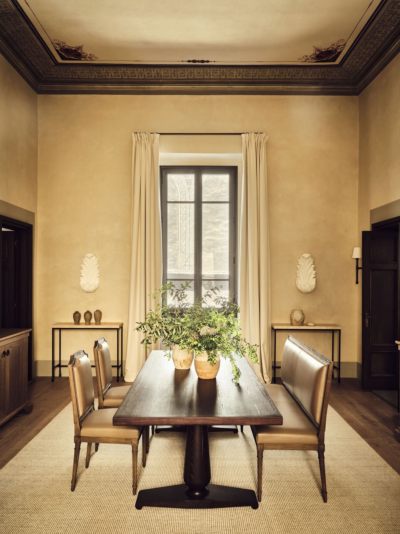 A dining room at La Réserve Firenze is framed by a painted Greek-key border on the ceiling and a pair of white acanthus ornaments on the walls. A dark oak pedestal table with leather-upholstered chairs and a bench stands on a woven rug, with olive branches in earthenware jars echoing the soft ochre palette. A dining room at La Réserve Firenze is framed by a painted Greek-key border on the ceiling and a pair of white acanthus ornaments on the walls. A dark oak pedestal table with leather-upholstered chairs and a bench stands on a woven rug, with olive branches in earthenware jars echoing the soft ochre palette.