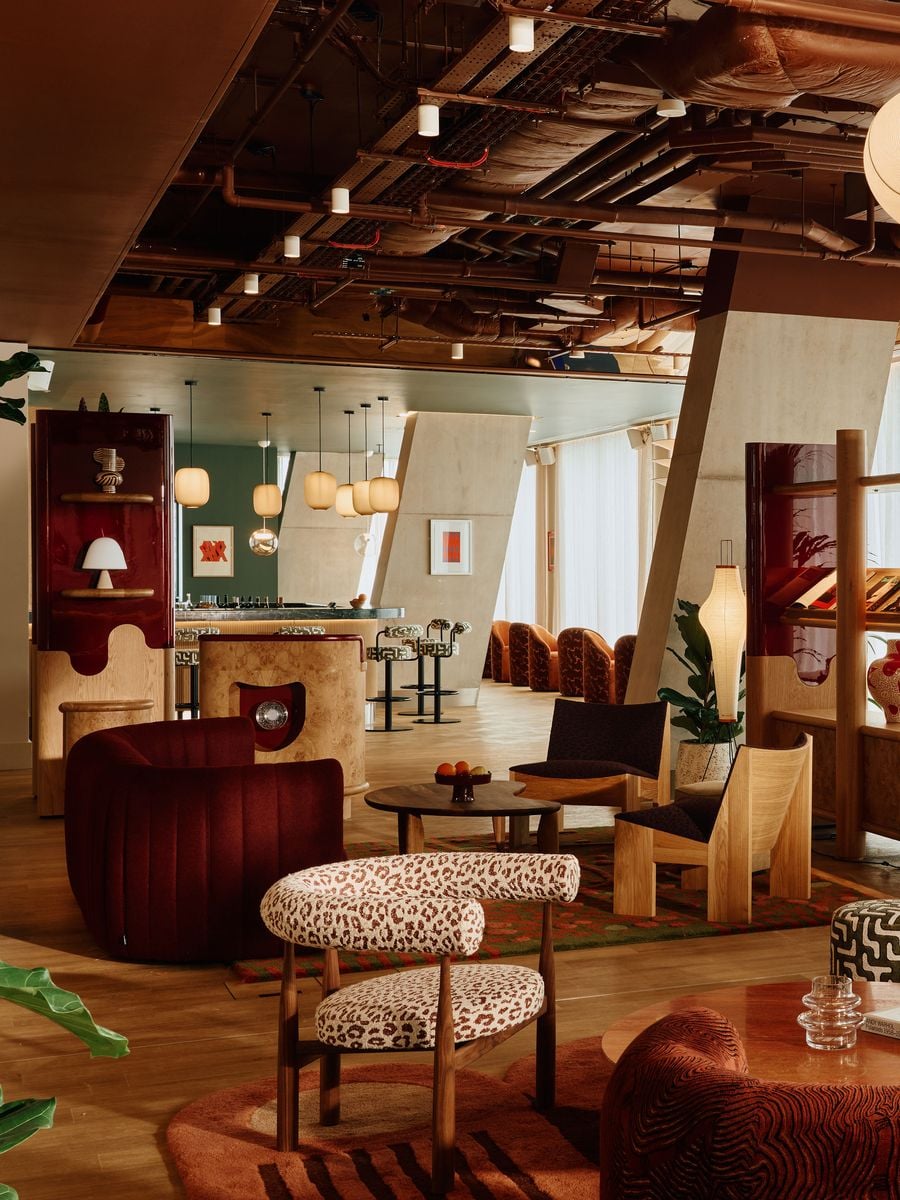 A wide view across the ground-floor lounge towards LUQA bar reveals the deliberate contrast between sculptural furniture and the building’s angular concrete structure. Burgundy seating, animal-print upholstery, patterned rugs and glossy bespoke shelving unfold beneath an exposed industrial ceiling, giving the open plan the character of interconnected cinematic sets. A wide view across the ground-floor lounge towards LUQA bar reveals the deliberate contrast between sculptural furniture and the building’s angular concrete structure. Burgundy seating, animal-print upholstery, patterned rugs and glossy bespoke shelving unfold beneath an exposed industrial ceiling, giving the open plan the character of interconnected cinematic sets.