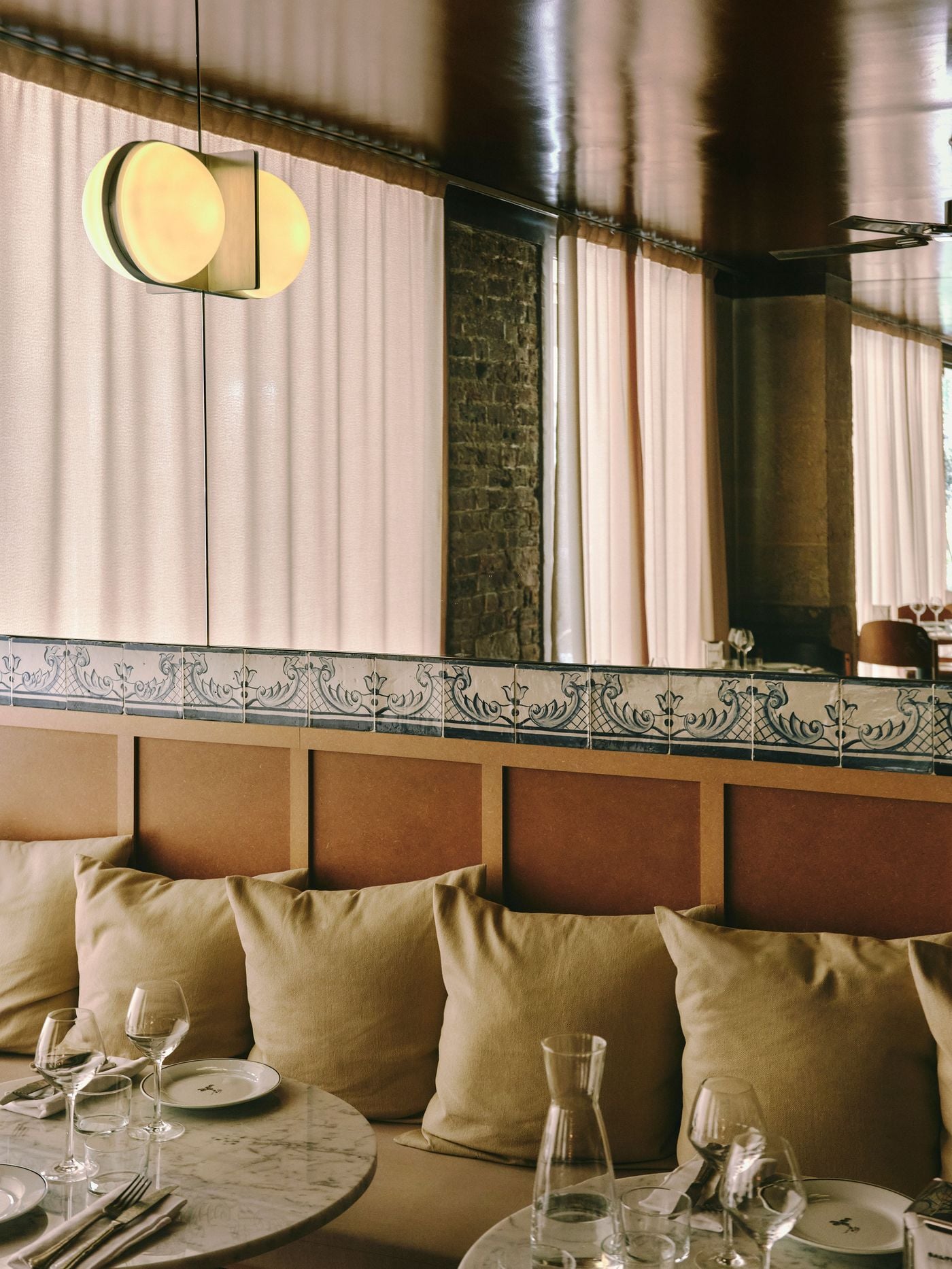 A frieze of Portuguese tiles with scrolling foliate motifs divides MDF-panelled seating from a mirrored upper wall at SaltoBar in Paris. The mirror reflects draped curtains and a sliver of brickwork. A double-disc pendant glows above plump linen cushions and marble tables laid with glassware and a water carafe. A frieze of Portuguese tiles with scrolling foliate motifs divides MDF-panelled seating from a mirrored upper wall at SaltoBar in Paris. The mirror reflects draped curtains and a sliver of brickwork. A double-disc pendant glows above plump linen cushions and marble tables laid with glassware and a water carafe.