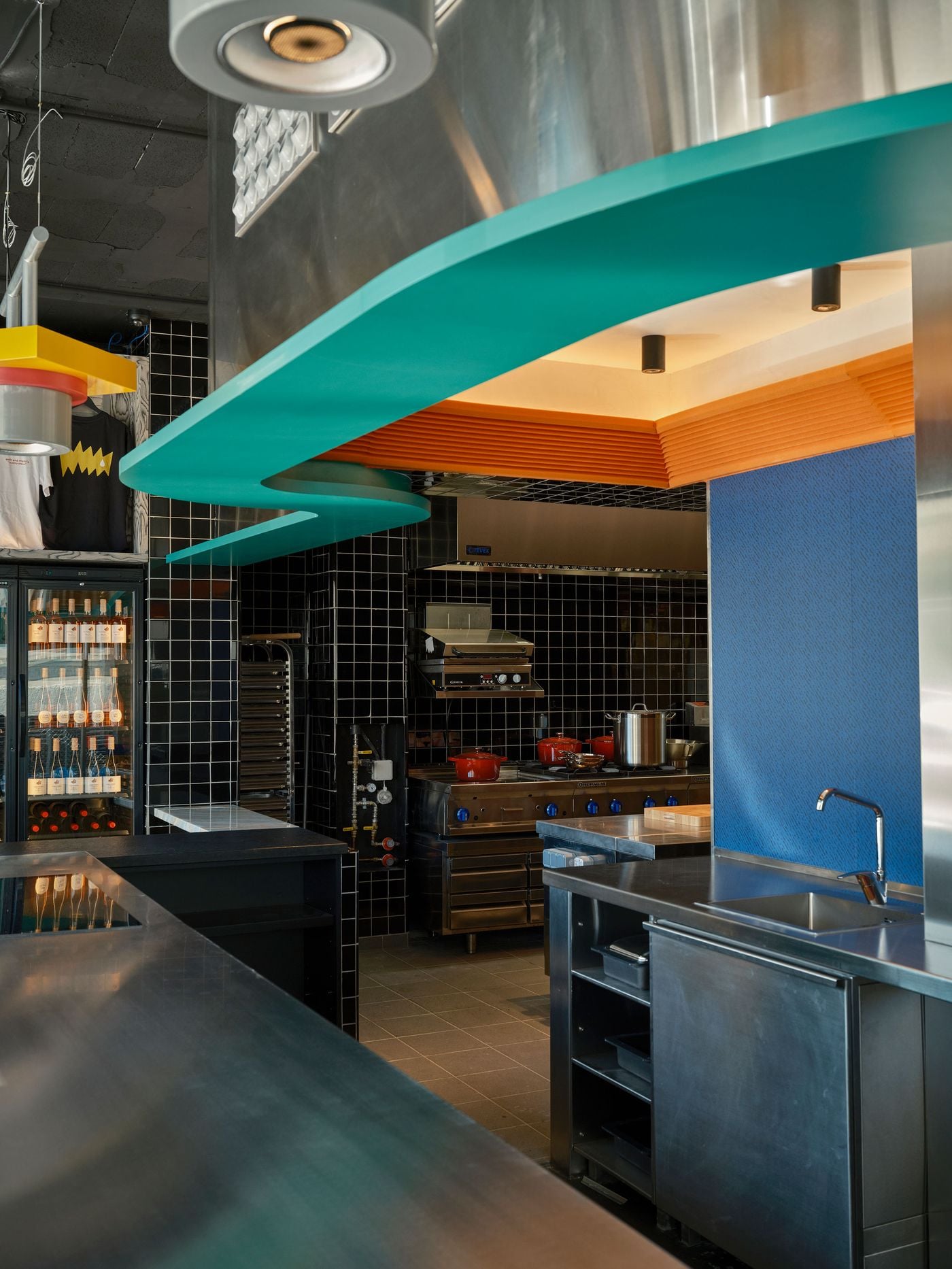 Beneath a sweeping teal overhang and an orange ribbed soffit, the open kitchen at Seth & Manni's reveals black-tiled walls, red enamel casseroles and stainless-steel ranges. A cobalt-blue panel and brushed-steel sink frame the right side. Wine fridges and branded T-shirts appear on the left. Beneath a sweeping teal overhang and an orange ribbed soffit, the open kitchen at Seth & Manni's reveals black-tiled walls, red enamel casseroles and stainless-steel ranges. A cobalt-blue panel and brushed-steel sink frame the right side. Wine fridges and branded T-shirts appear on the left.