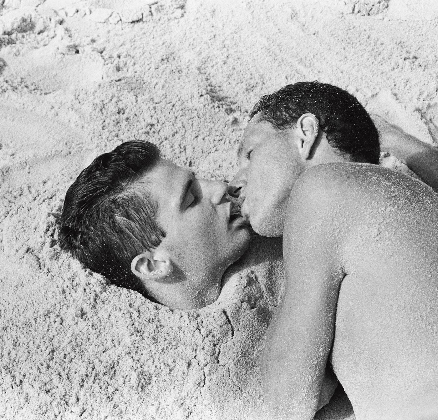 Two men kiss on a beach, one half-buried in the sand, his eyes closed, as the other leans over him, grains glittering on his shoulder and cheek. The fine texture of the sand and the soft tonal range of this black-and-white photograph from Don't Worry Boys lend the moment a sculptural stillness. Two men kiss on a beach, one half-buried in the sand, his eyes closed, as the other leans over him, grains glittering on his shoulder and cheek. The fine texture of the sand and the soft tonal range of this black-and-white photograph from Don't Worry Boys lend the moment a sculptural stillness.