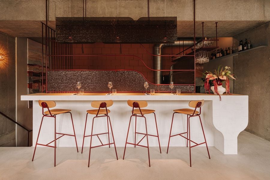 The upstairs dining counter at WIN wine bar in Warsaw pairs a sculptural white-tiled base with a warm timber top and slender burgundy-framed stools. Behind it, undulating metalwork and textured glass panels form a semi-transparent partition, while a suspended canopy adds another layer of texture against the exposed concrete ceiling.