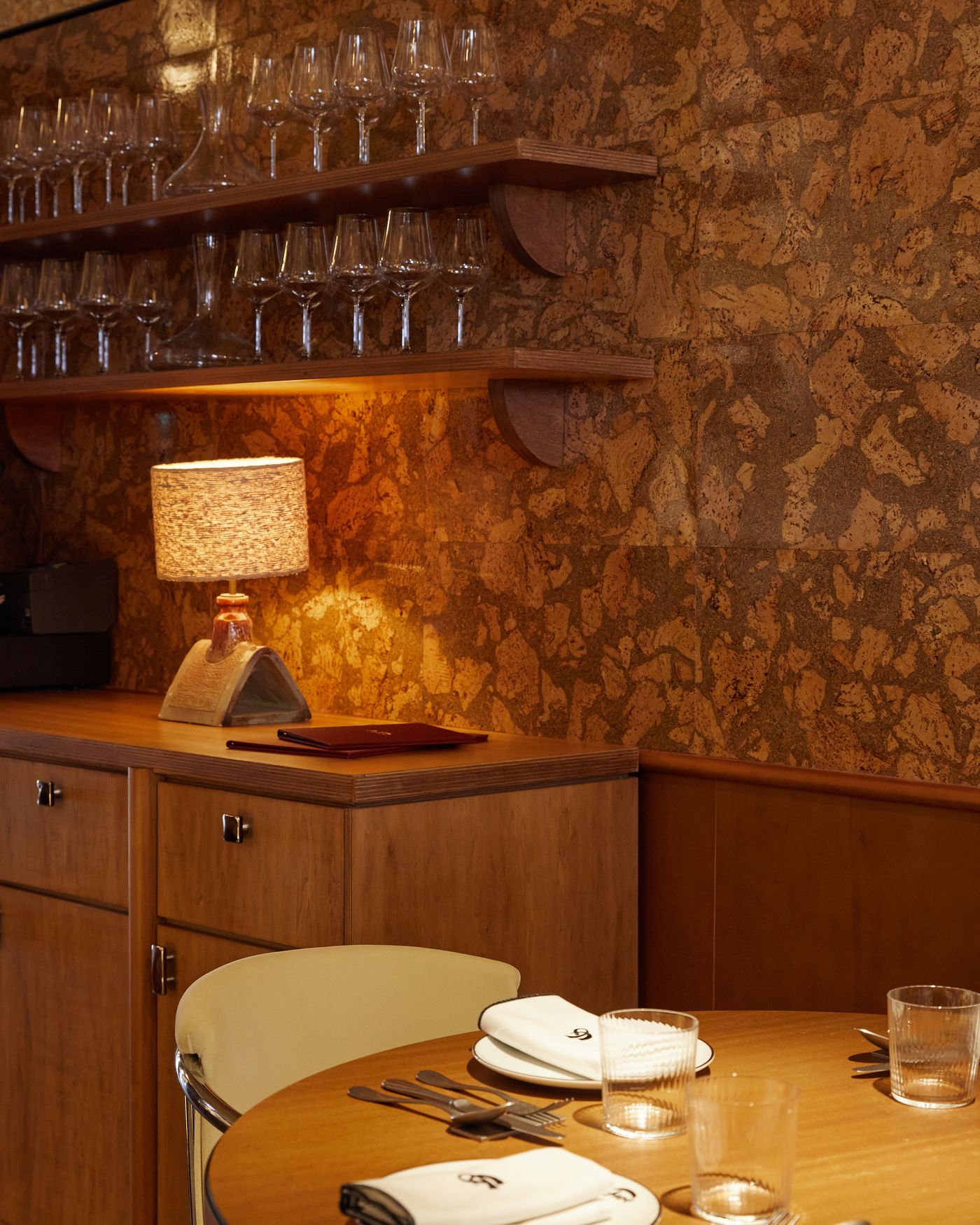 A dining area defined by rich textures and warm tones. The wall, clad in a unique cork-like material, is complemented by a wooden sideboard and shelves holding delicate glassware. A small vintage-style lamp adds an intimate glow, highlighting the space's tactile quality.