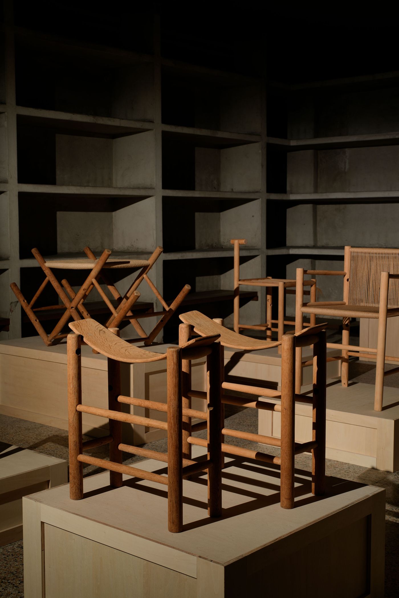 A collection of wooden chairs displayed on light-colored pedestals in a dimly lit space, showcasing various designs and textures.