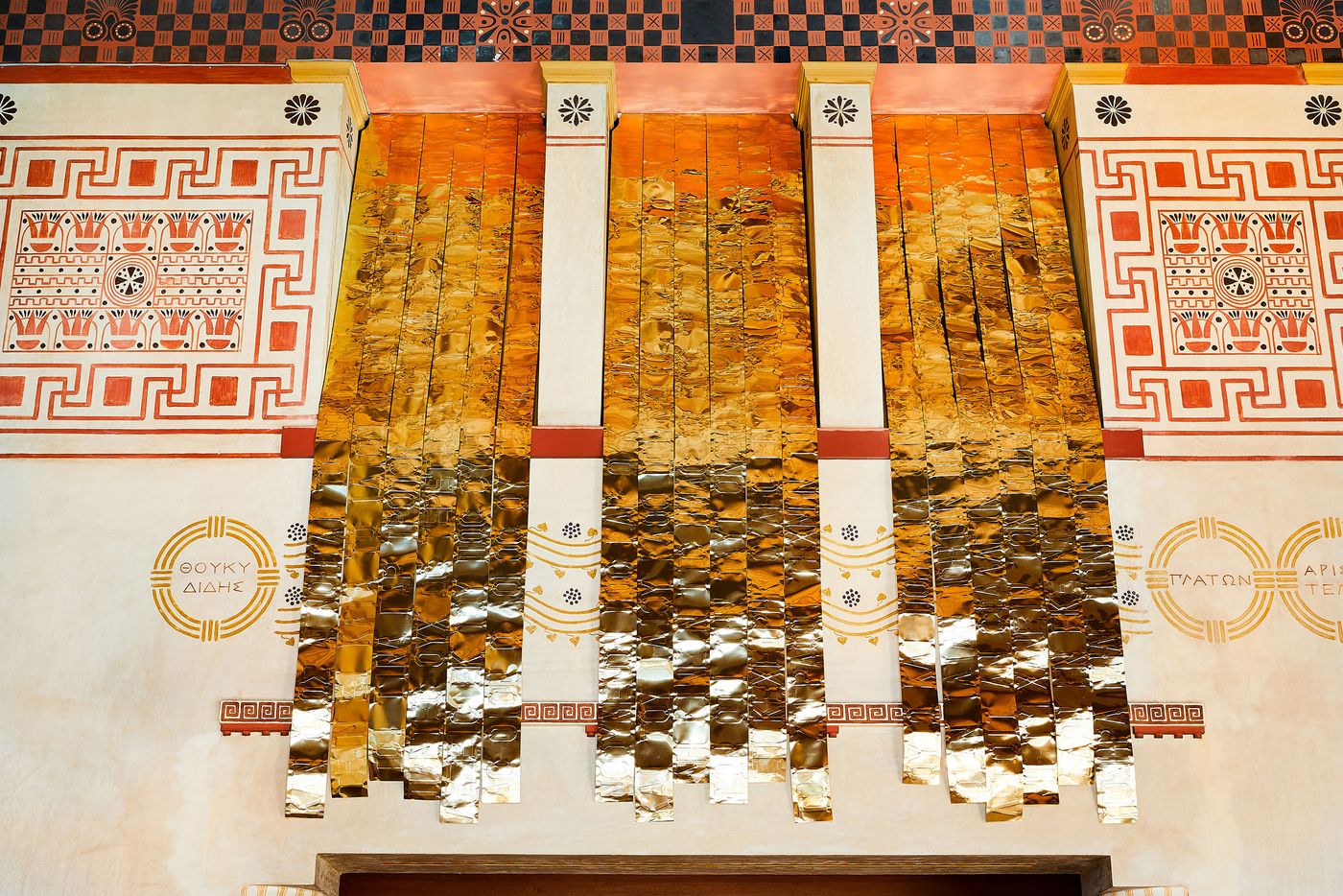 A shot of the upper wall in a Grecian-style room. The wall is adorned with red and black geometric friezes. Three vertical panels of rippling golden metal, part of a Gabriel Leger installation, hang from the ceiling, their reflective surfaces distorting the patterns on the walls and adding a modern, reflective layer to the traditional setting. A shot of the upper wall in a Grecian-style room. The wall is adorned with red and black geometric friezes. Three vertical panels of rippling golden metal, part of a Gabriel Leger installation, hang from the ceiling, their reflective surfaces distorting the patterns on the walls and adding a modern, reflective layer to the traditional setting.