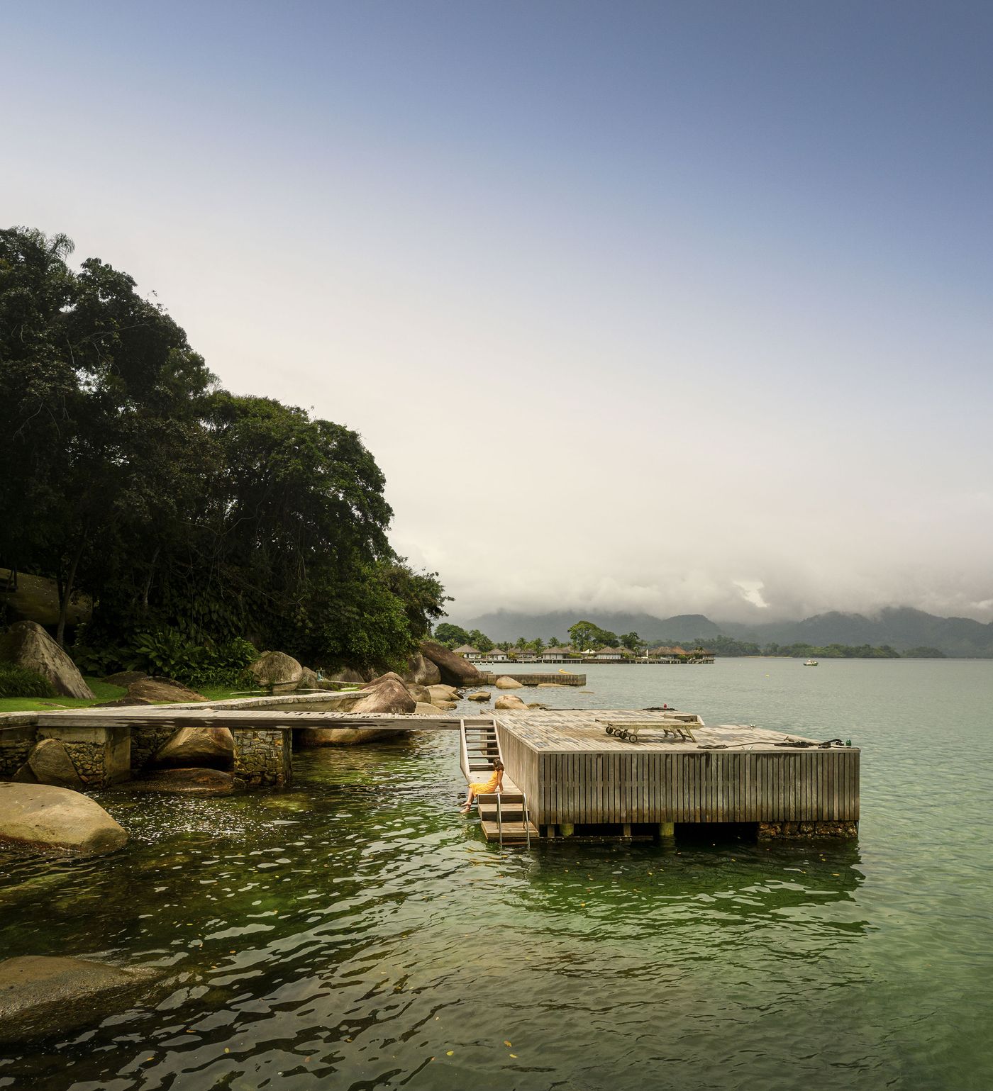 A sturdy wooden pier with a small swimming platform extends into the calm, green-tinted waters, complementing the rocky, tree-lined coastline. The natural textures of wood and stone are prominent, creating an authentic, rustic-modern transition from land to sea under a soft, overcast sky.