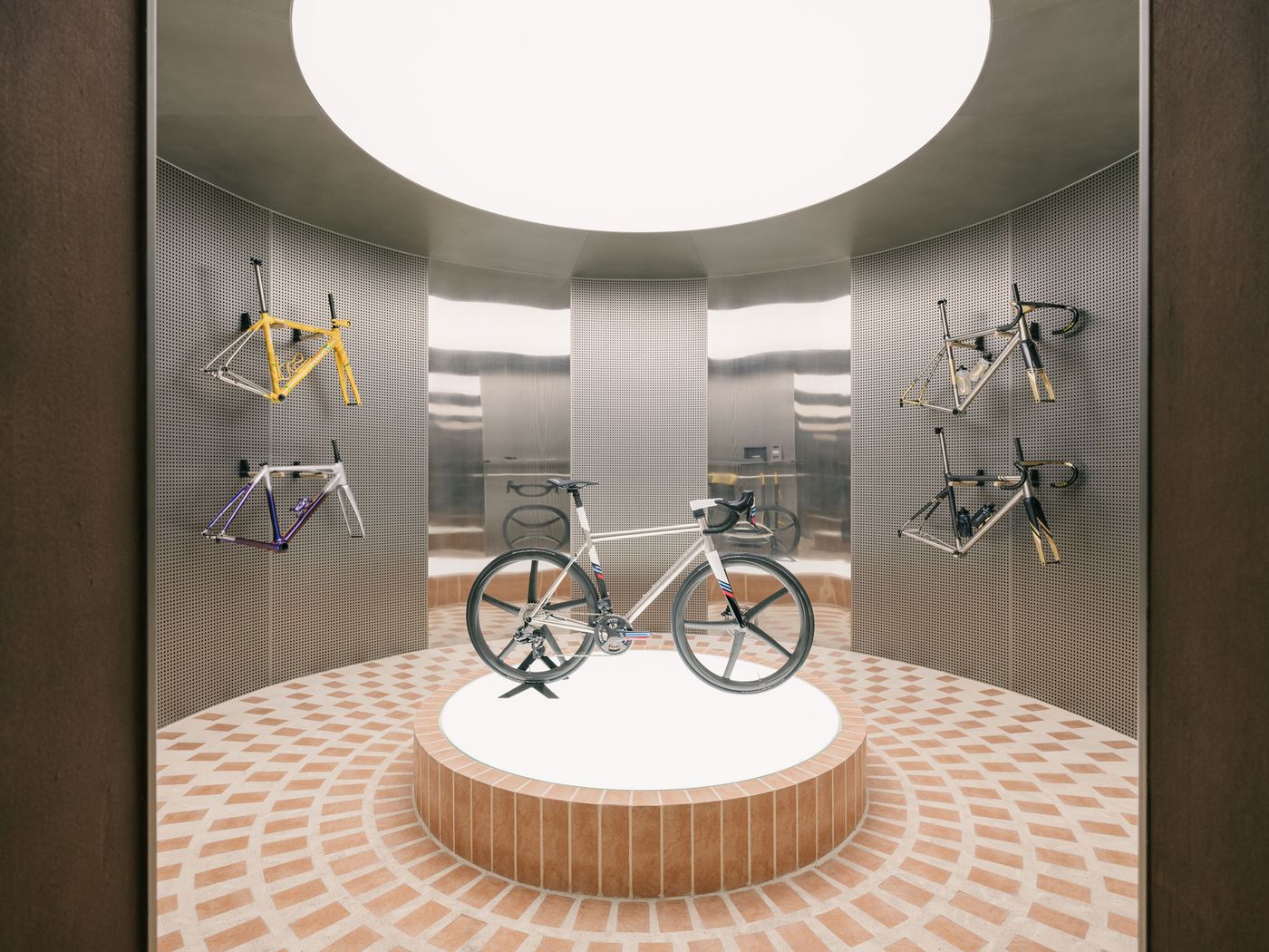 A focused, circular display acts as a modern shrine for a bicycle, dramatically lit from an overhead oculus. The surrounding walls are clad in polished, perforated metal, reflecting the light and providing a textured backdrop for hanging bicycle frames in vibrant colors, all set against a warm, radially-patterned terracotta tile floor. A focused, circular display acts as a modern shrine for a bicycle, dramatically lit from an overhead oculus. The surrounding walls are clad in polished, perforated metal, reflecting the light and providing a textured backdrop for hanging bicycle frames in vibrant colors, all set against a warm, radially-patterned terracotta tile floor.