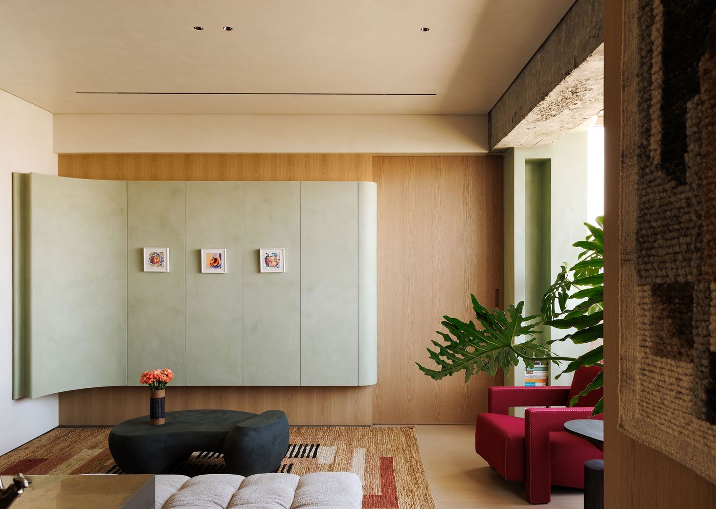 A minimalist yet inviting living area designed by YSP Design, featuring a striking pale green curved wall unit with subtle framed art. The warm wood paneling creates a serene backdrop, complemented by a large green plant and a bold red armchair. A sculptural dark gray coffee table and a textured rug in earthy tones complete the sophisticated, art-infused atmosphere. A minimalist yet inviting living area designed by YSP Design, featuring a striking pale green curved wall unit with subtle framed art. The warm wood paneling creates a serene backdrop, complemented by a large green plant and a bold red armchair. A sculptural dark gray coffee table and a textured rug in earthy tones complete the sophisticated, art-infused atmosphere.