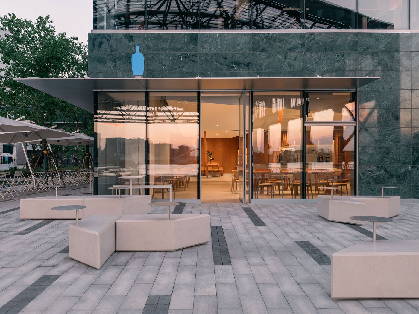 The glazed exterior of Blue Bottle Coffee reflects the riverside cityscape as sculptural concrete seating clusters animate the forecourt, blending the café’s artisanal ethos with Shanghai’s modern urban rhythm.