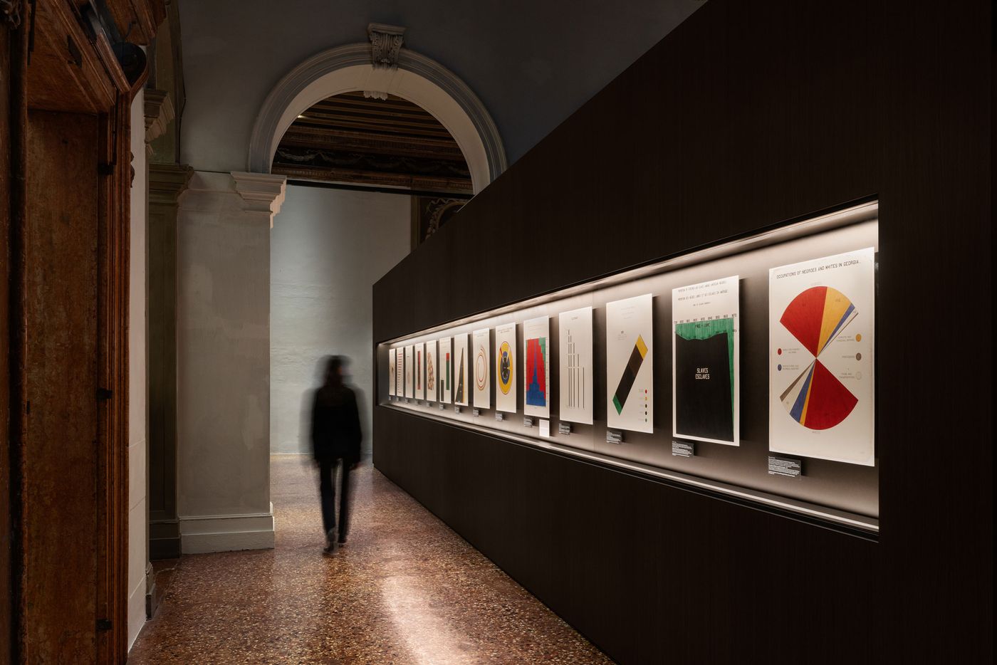 A visitor walks past a gallery wall displaying various colorful infographics in a softly lit exhibition space. A visitor walks past a gallery wall displaying various colorful infographics in a softly lit exhibition space.