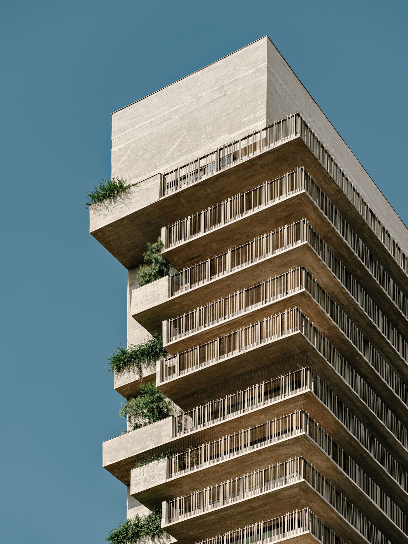 An upward angle showcasing the top floors of the tower, revealing the striking cantilevered concrete balconies and a refined system of vertical metal balustrades. Greenery emerges from some terraces, subtly integrating nature into the minimalist, industrial aesthetic against a clear blue sky. An upward angle showcasing the top floors of the tower, revealing the striking cantilevered concrete balconies and a refined system of vertical metal balustrades. Greenery emerges from some terraces, subtly integrating nature into the minimalist, industrial aesthetic against a clear blue sky.