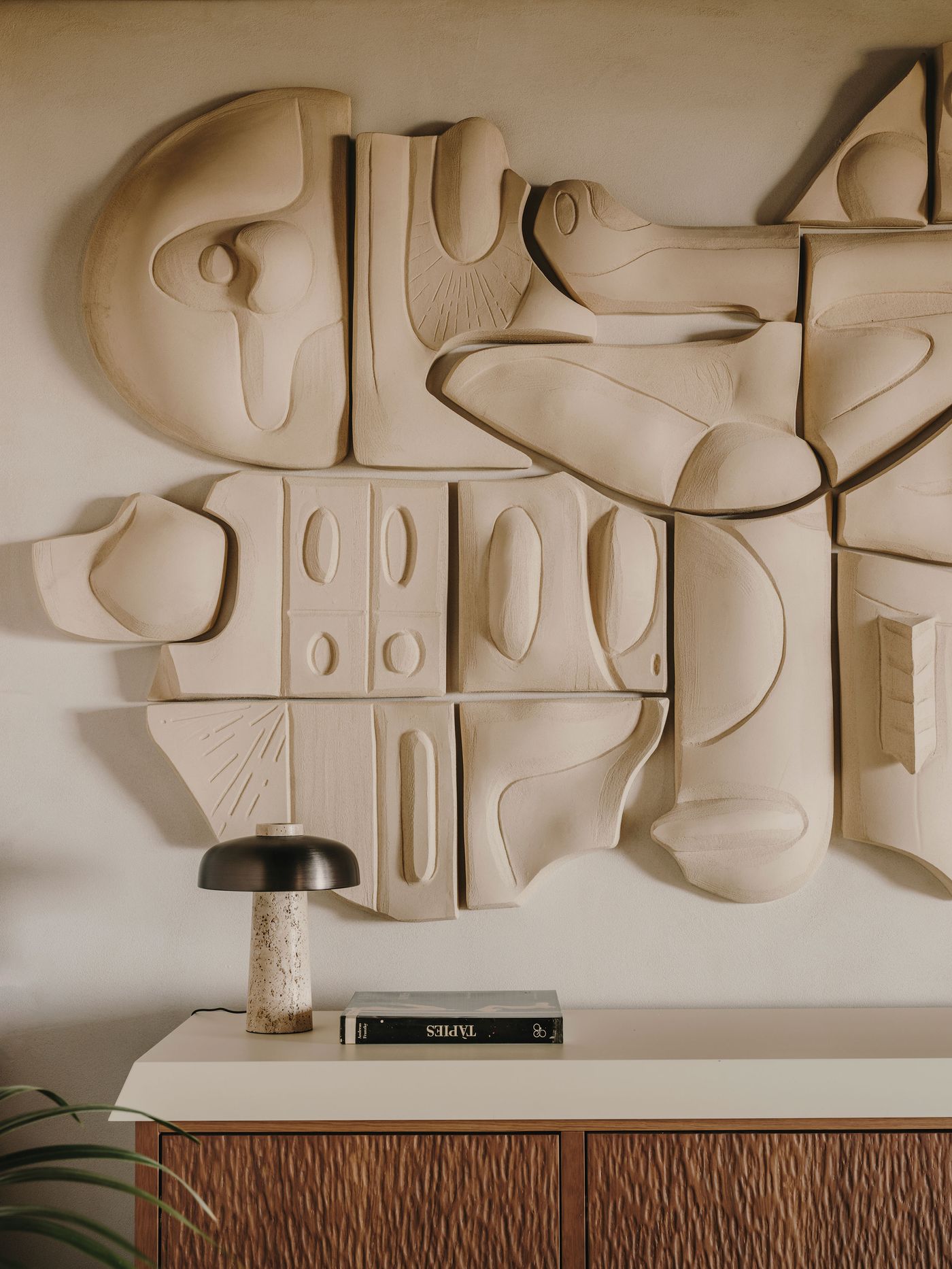 A textured, abstract wall sculpture hangs above a modern side table with a lamp and a book, creating a stylish, contemporary interior.
