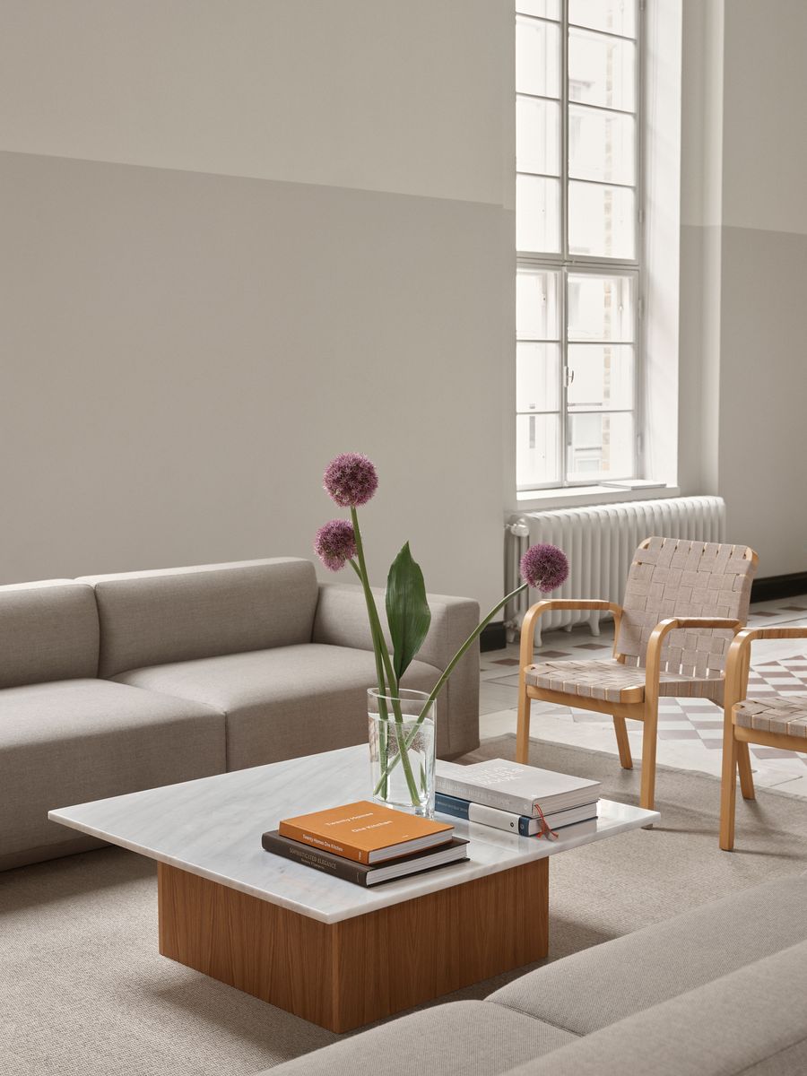 A minimalist lounge area features a light grey sofa and two woven-back chairs, centered around a multi-level coffee table with flowers and books. A large window provides natural light, enhancing the tranquil and uncluttered aesthetic of the space. A minimalist lounge area features a light grey sofa and two woven-back chairs, centered around a multi-level coffee table with flowers and books. A large window provides natural light, enhancing the tranquil and uncluttered aesthetic of the space.