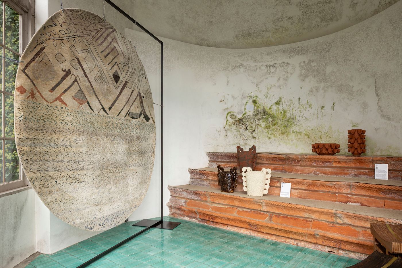 An interior exhibition space features a large, circular textile artwork with abstract patterns of brown lines on a textured, neutral background, displayed on a metal stand. Adjacent to it, terracotta-colored ceramic vessels with unique, organic shapes are arranged on stepped, brick-like platforms. The vibrant turquoise tiled floor and the subtly distressed walls add to the room's artistic atmosphere, suggesting a fusion of craft and design.