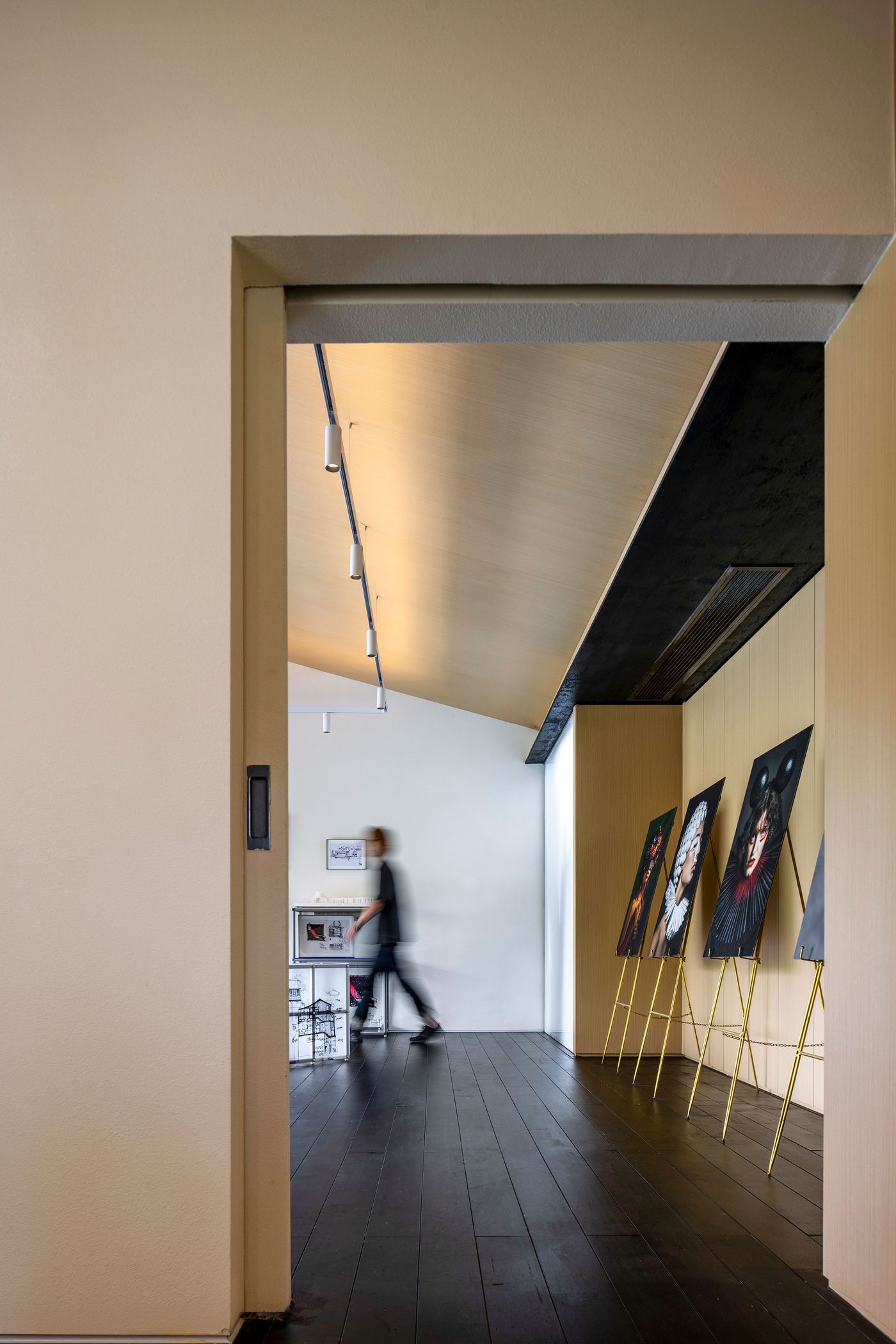 An art gallery-like corridor within Wutopia Lab's Lake House features a striking composition of light and shadow. Dark wooden floors lead the eye past a series of modern artworks displayed on minimalist gold easels, creating a dynamic visual flow. A person in motion adds a sense of life to the space, which is illuminated by track lighting against a light-colored, angled ceiling. An art gallery-like corridor within Wutopia Lab's Lake House features a striking composition of light and shadow. Dark wooden floors lead the eye past a series of modern artworks displayed on minimalist gold easels, creating a dynamic visual flow. A person in motion adds a sense of life to the space, which is illuminated by track lighting against a light-colored, angled ceiling.