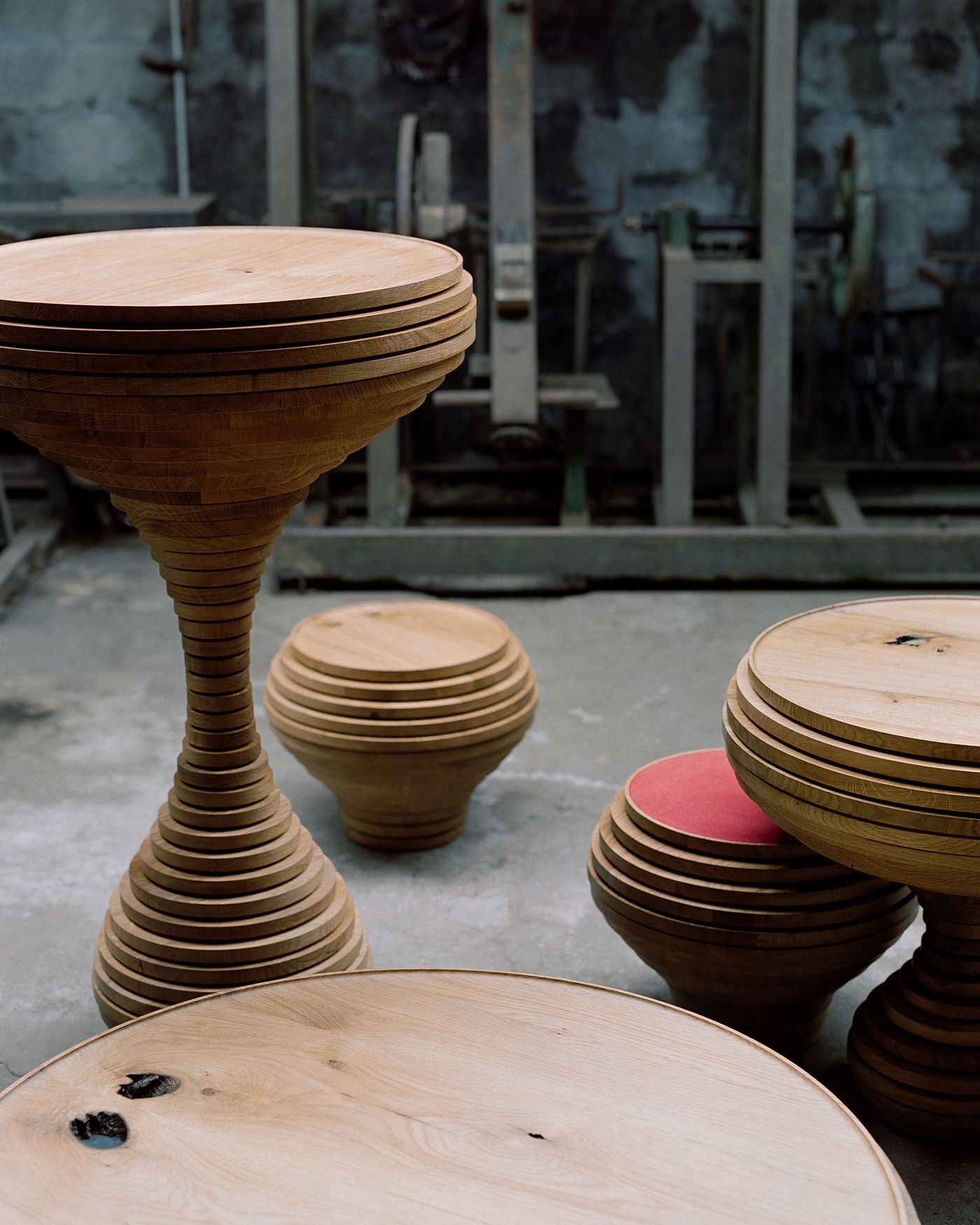 Sculptural wooden vessels display masterful lathe work, their layered construction creating organic forms reminiscent of pottery. The industrial workshop setting with concrete floors provides a raw backdrop, highlighting the contrast between refined craftsmanship and utilitarian space in contemporary furniture design.