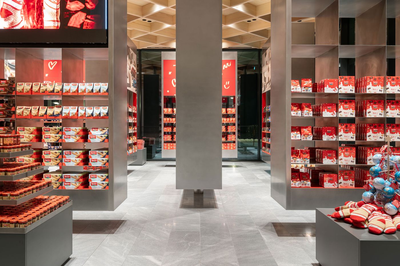 Monumental retail displays reach toward a geometrically coffered ceiling, creating cathedral-like proportions for product presentation. The systematic organization of colorful merchandise against neutral architecture transforms shopping into cultural experience, where commerce meets contemporary gallery aesthetic through precise spatial design.