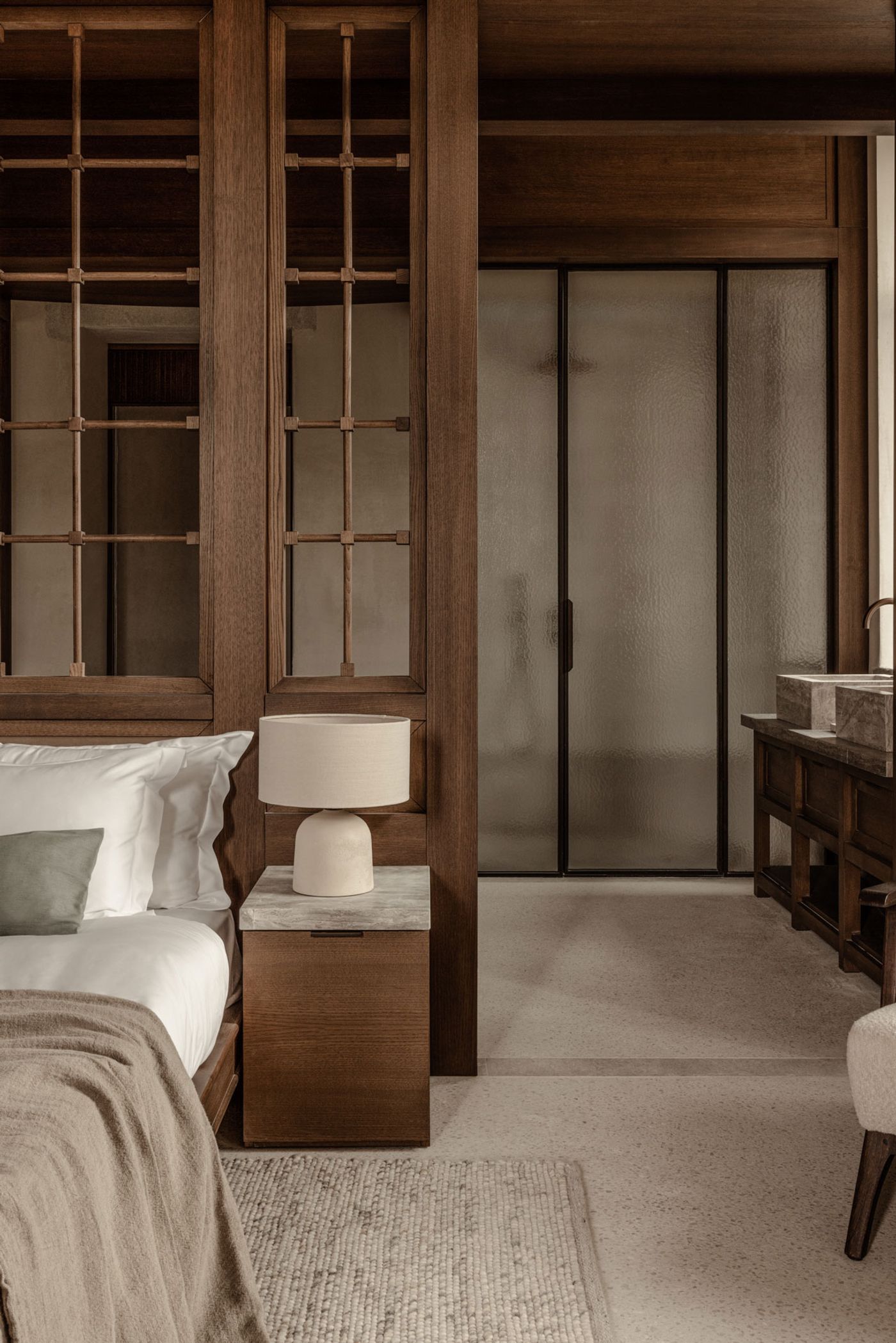 This intimate interior view captures a blend of traditional craftsmanship and modern design. Dark timber joinery with delicate lattice work defines the sleeping area, contrasting with the frosted glass sliding doors of the bathroom, which features a stone vanity and vessel sinks, embodying a sophisticated Japandi influence. This intimate interior view captures a blend of traditional craftsmanship and modern design. Dark timber joinery with delicate lattice work defines the sleeping area, contrasting with the frosted glass sliding doors of the bathroom, which features a stone vanity and vessel sinks, embodying a sophisticated Japandi influence.