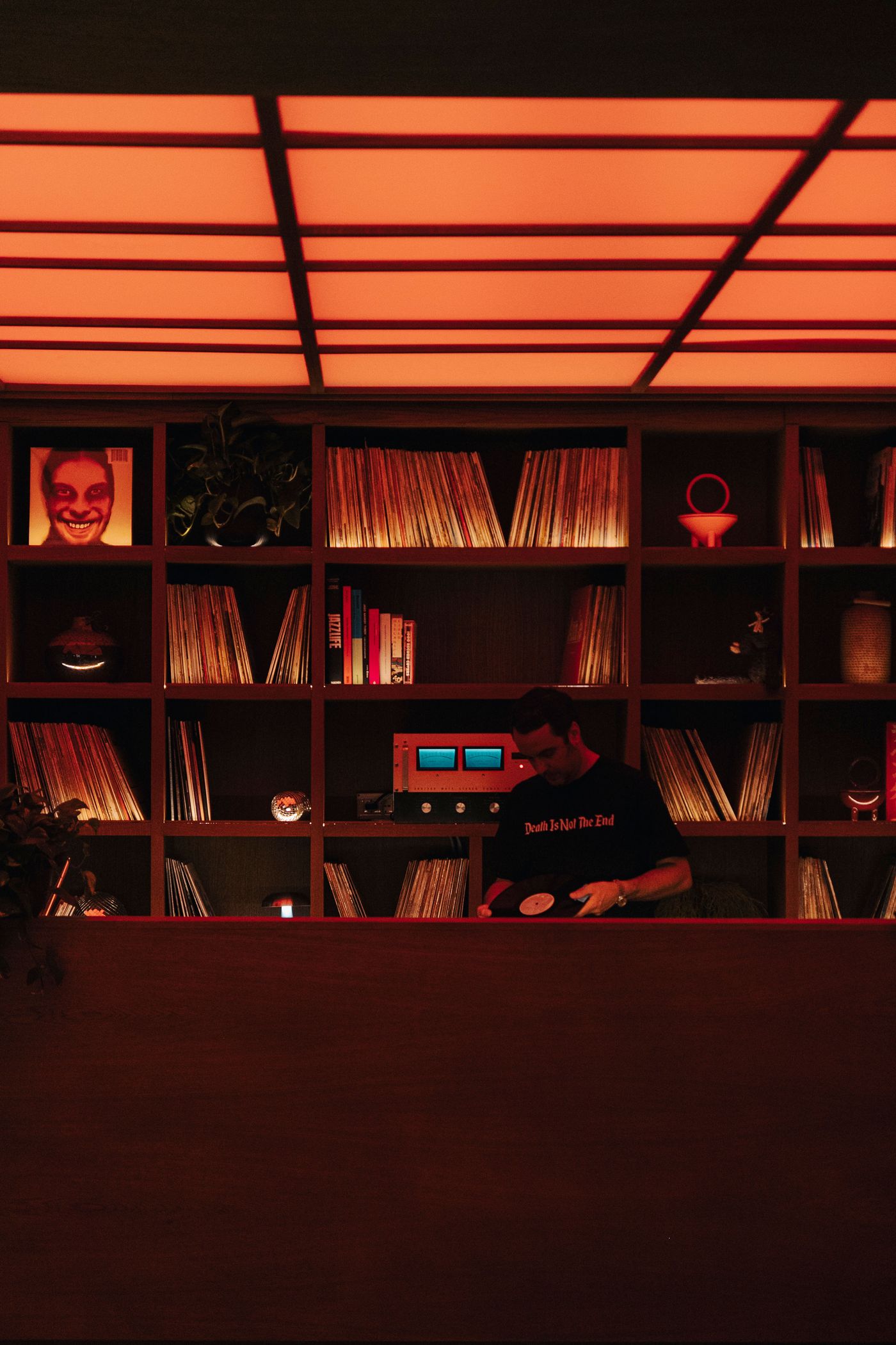 A dimly-lit room filled with vinyl records and a retro sound system, highlighted by warm orange lighting. A person holds a record.