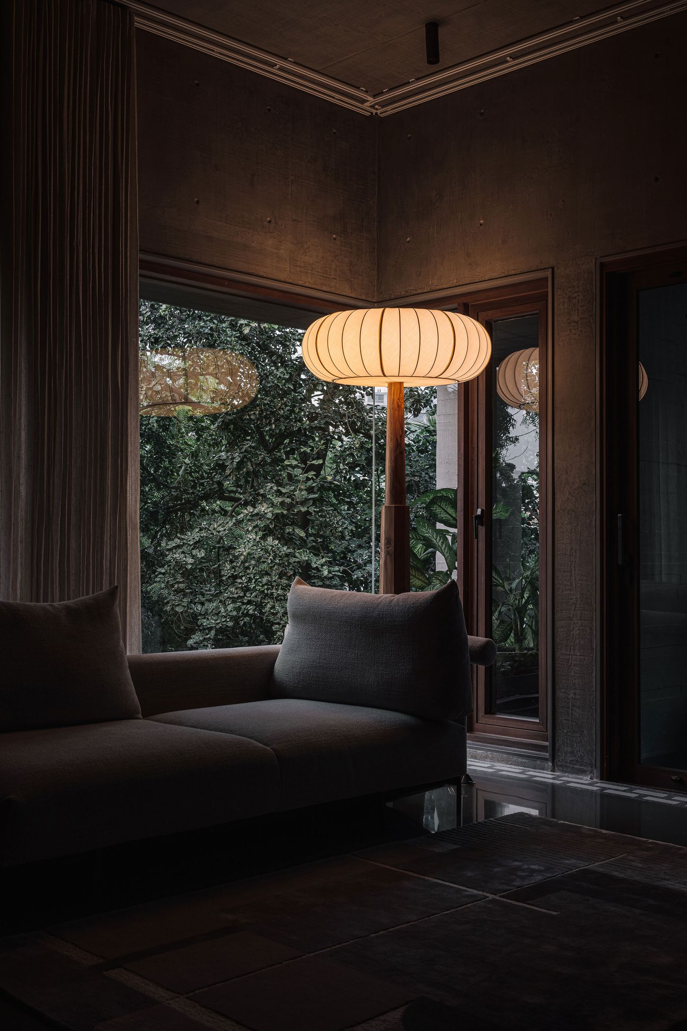 A dimly lit, inviting living space, where the soft glow of a spherical, ribbed pendant lamp illuminates a comfortable, dark grey sofa. Large windows frame a verdant outdoor landscape, bringing nature indoors. The raw concrete walls and dark floor create a moody, sophisticated atmosphere, balanced by the warmth of natural light.