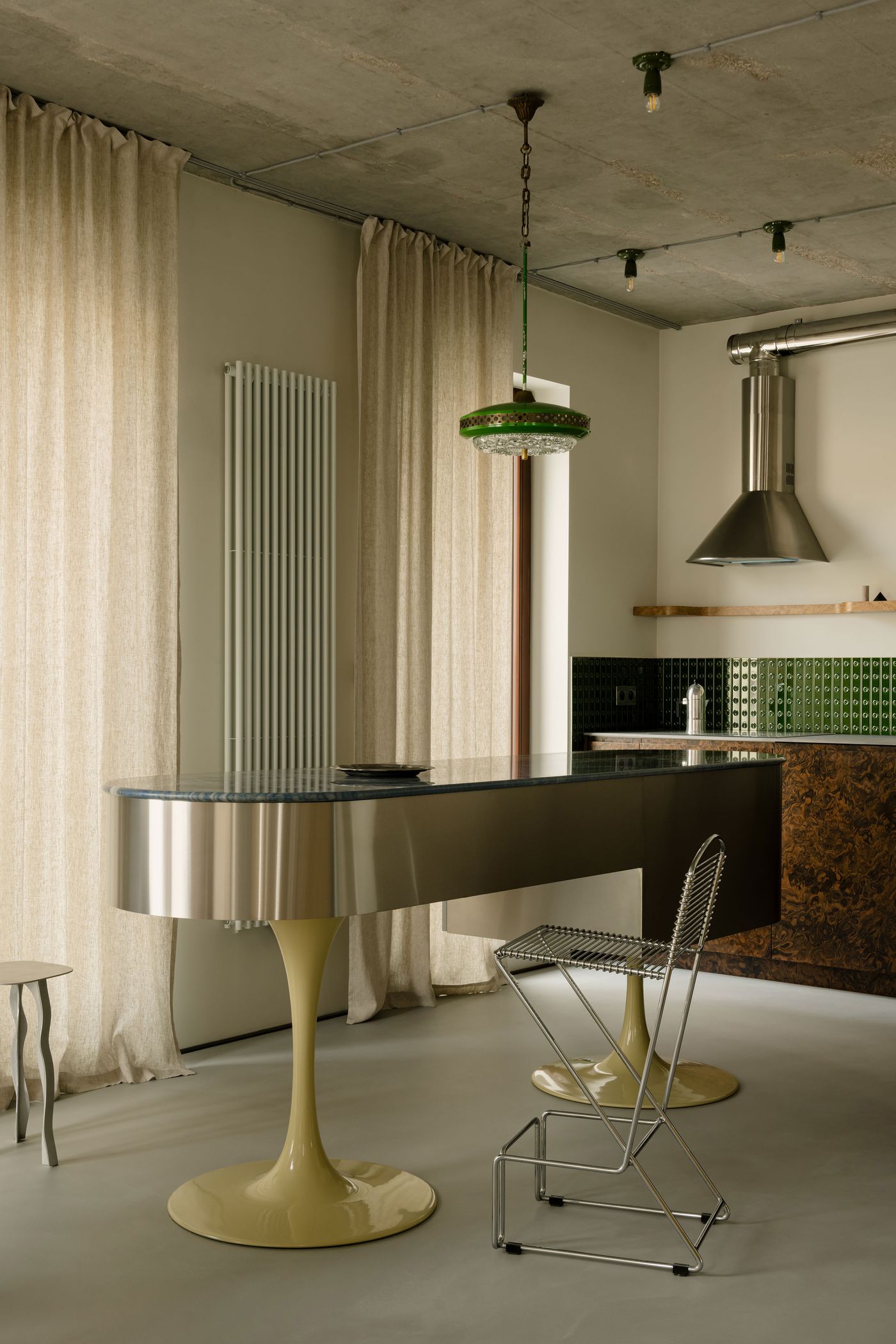 Curtains filter in natural light, softening the contrast between the kitchen’s reflective surfaces and raw concrete ceiling. The whimsical silhouette of a steel barstool adds a functional sculptural element. Curtains filter in natural light, softening the contrast between the kitchen’s reflective surfaces and raw concrete ceiling. The whimsical silhouette of a steel barstool adds a functional sculptural element.