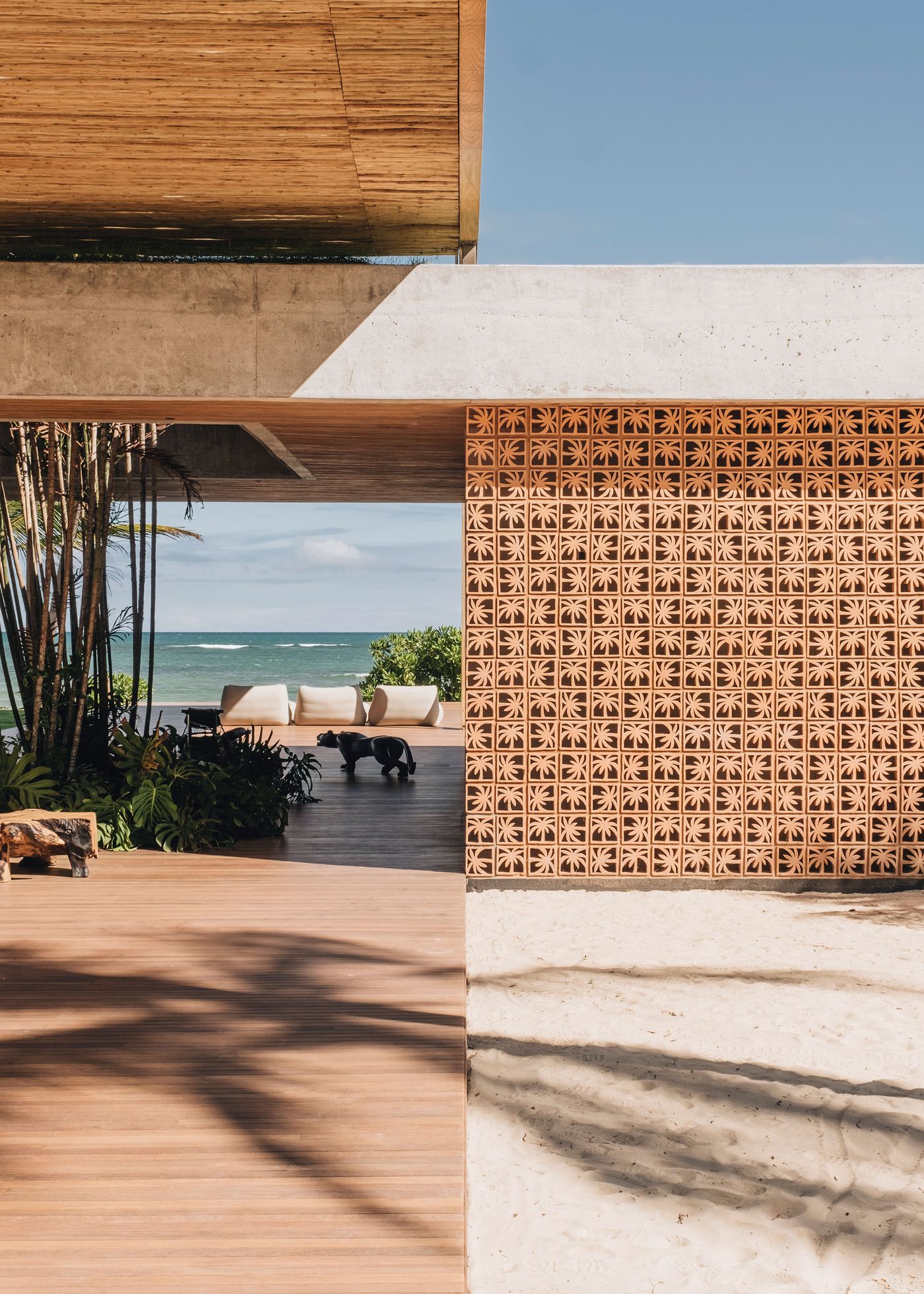 A modern open space with a terracotta block wall, wooden flooring, and a coastal view featuring lush plants and a playful black dog.