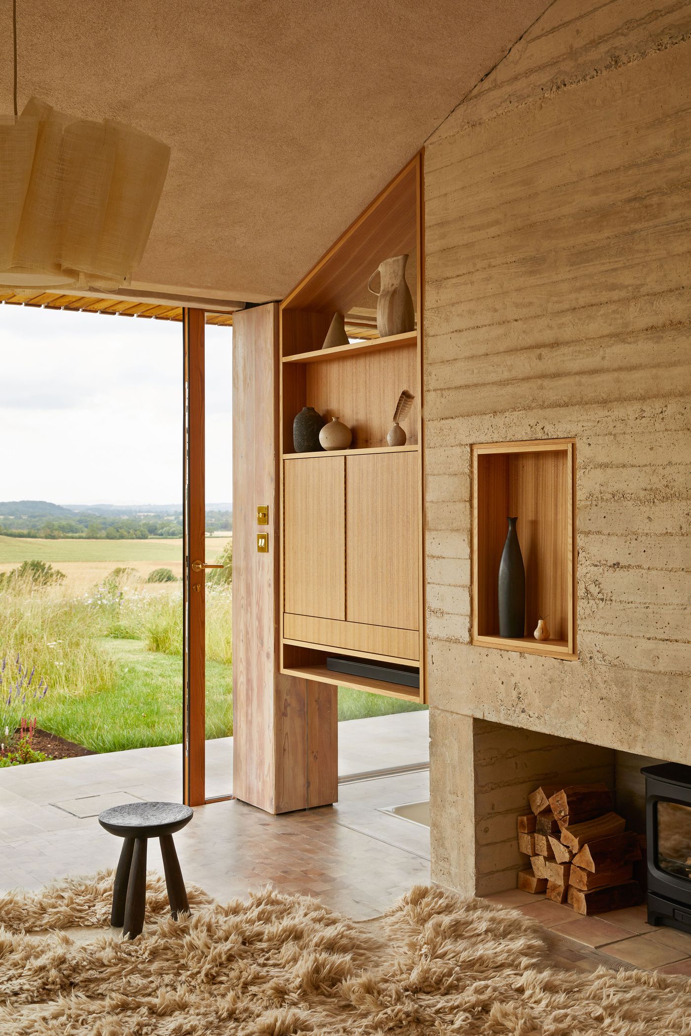 The interior of The Maker's Barn by Hutch Design reveals a sophisticated blend of natural materials. A textured rammed earth wall features built-in wooden shelving and a niche for art, complementing a wood-burning stove with stacked logs. The warm, inviting ambiance is enhanced by a plush sheepskin rug, a dark minimalist stool, and the expansive natural light from the open doorway to the wild outdoors. The interior of The Maker's Barn by Hutch Design reveals a sophisticated blend of natural materials. A textured rammed earth wall features built-in wooden shelving and a niche for art, complementing a wood-burning stove with stacked logs. The warm, inviting ambiance is enhanced by a plush sheepskin rug, a dark minimalist stool, and the expansive natural light from the open doorway to the wild outdoors.